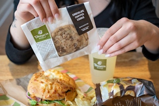 A person holding a Chocolate Chipper cookie next to a Panera meal.