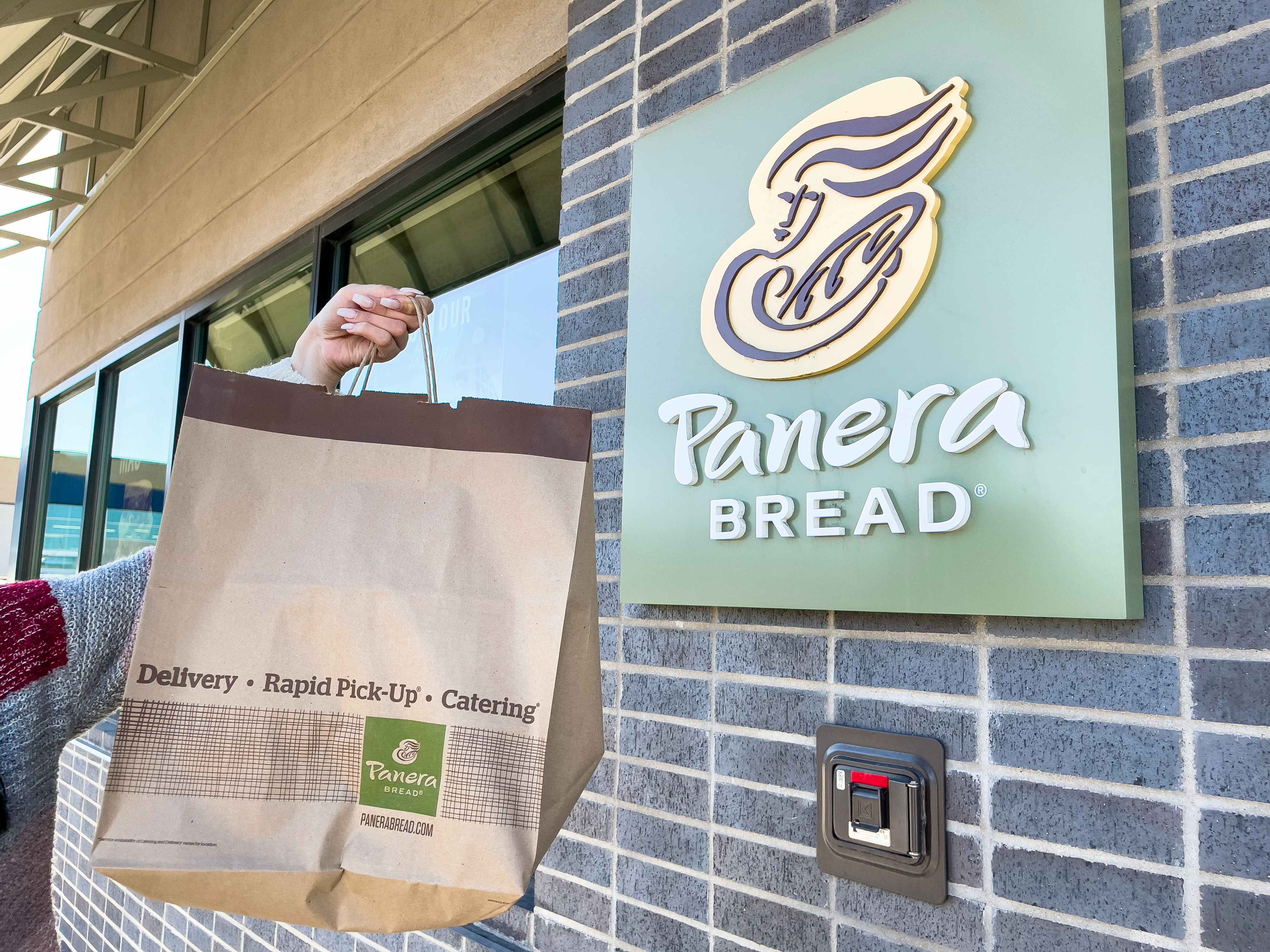 A person holding a paper bag filled with food in front of a Panera restaurant