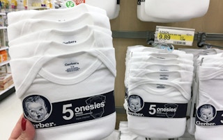 A person holding a pack of Gerber onesies in front of other packs of onesies hanging on a store display.
