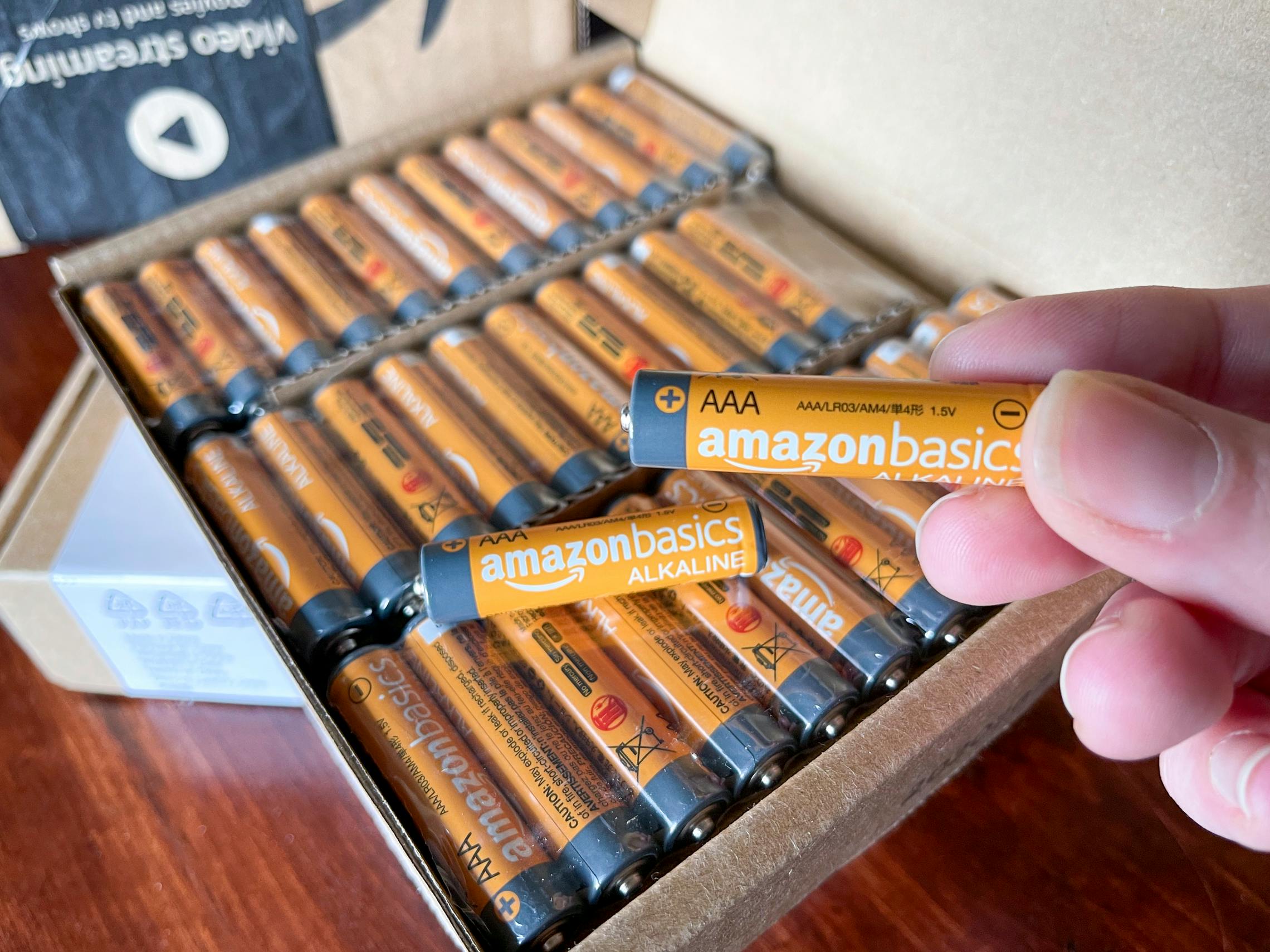 A person holding up an orange amazon basics battery with an open box of batteries in the background.