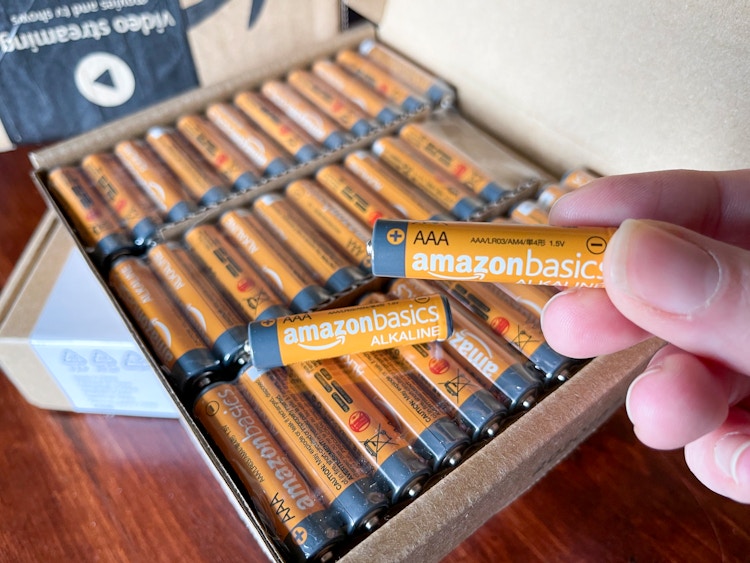 A person holding up an orange amazon basics battery with an open box of batteries in the background.