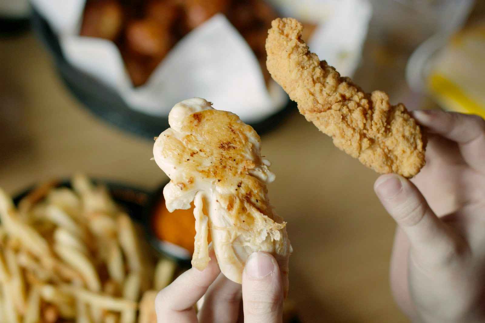 A hand holding on breaded chicken tender and the other holding one non-breaded chicken tender above a table with a basket of wings and a basket of fries..