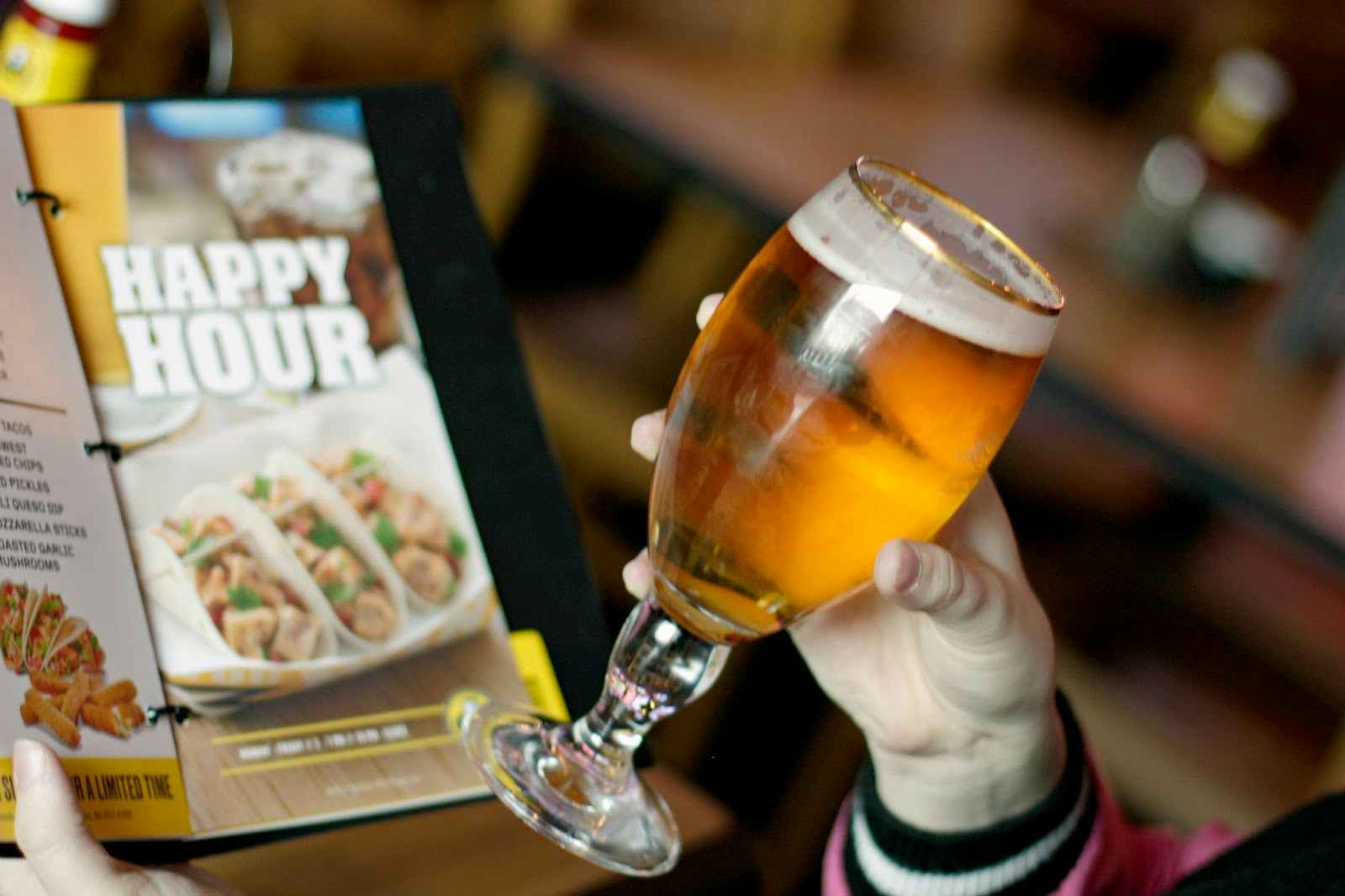 A hand holding a glass of beer with a Happy Hour menu in the background.