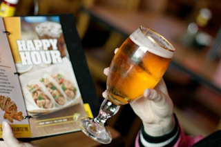 A hand holding a glass of beer with a Happy Hour menu in the background.