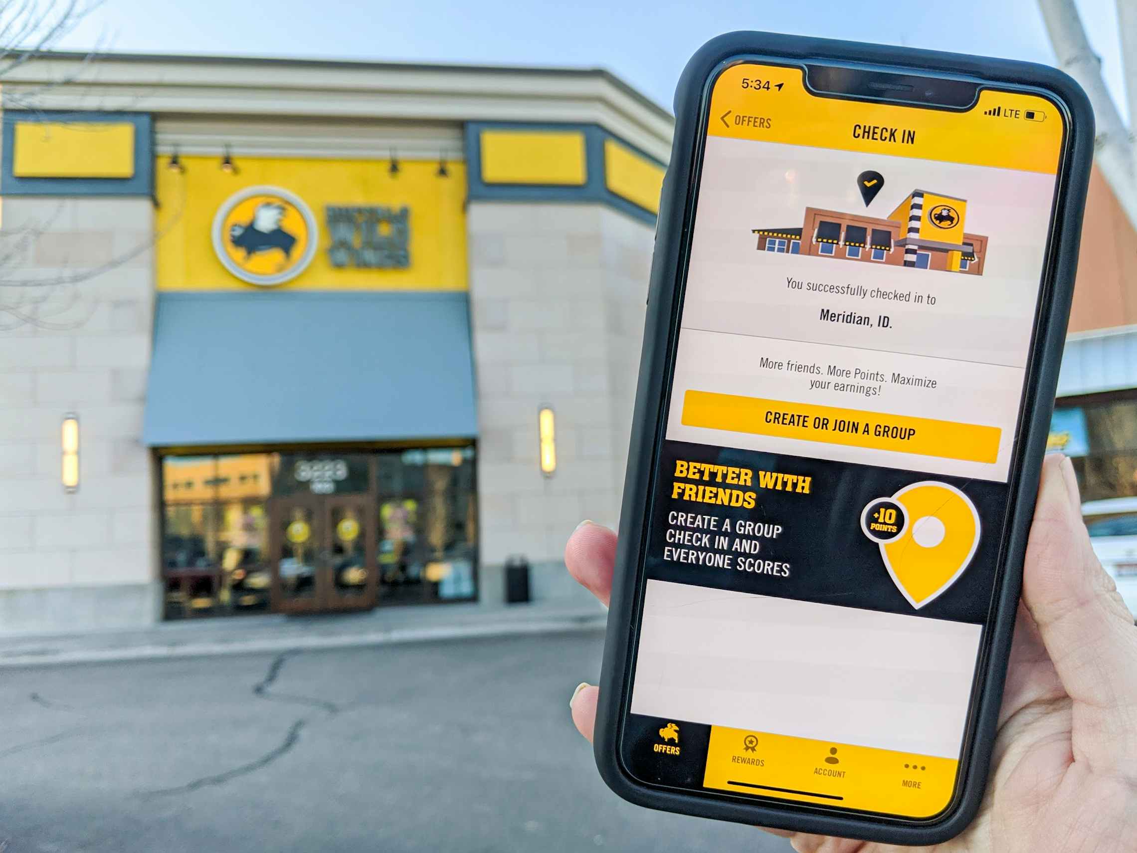 A person holding their iPhone up showing the check-in page of the Buffalo Wild Wings mobile app in front of a Buffalo Wild Wings storefront.