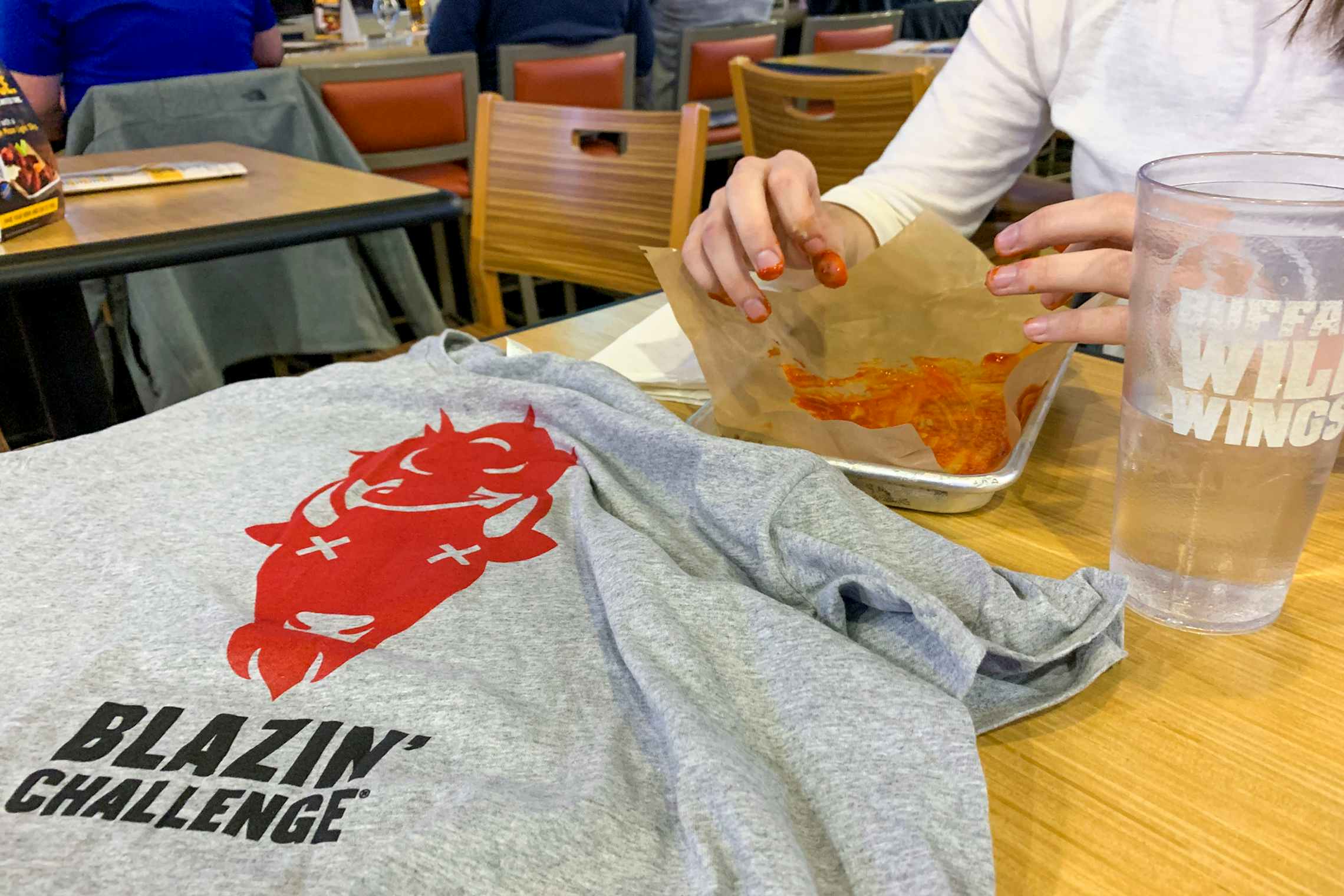 A woman sitting in a Buffalo Wild Wings restaurant with her fingers covered in buffalo sauce over an empty platter. There is a cup of water on the table next to her and a t-shirt with a graphic of a buffalo with X's for eyes that says 