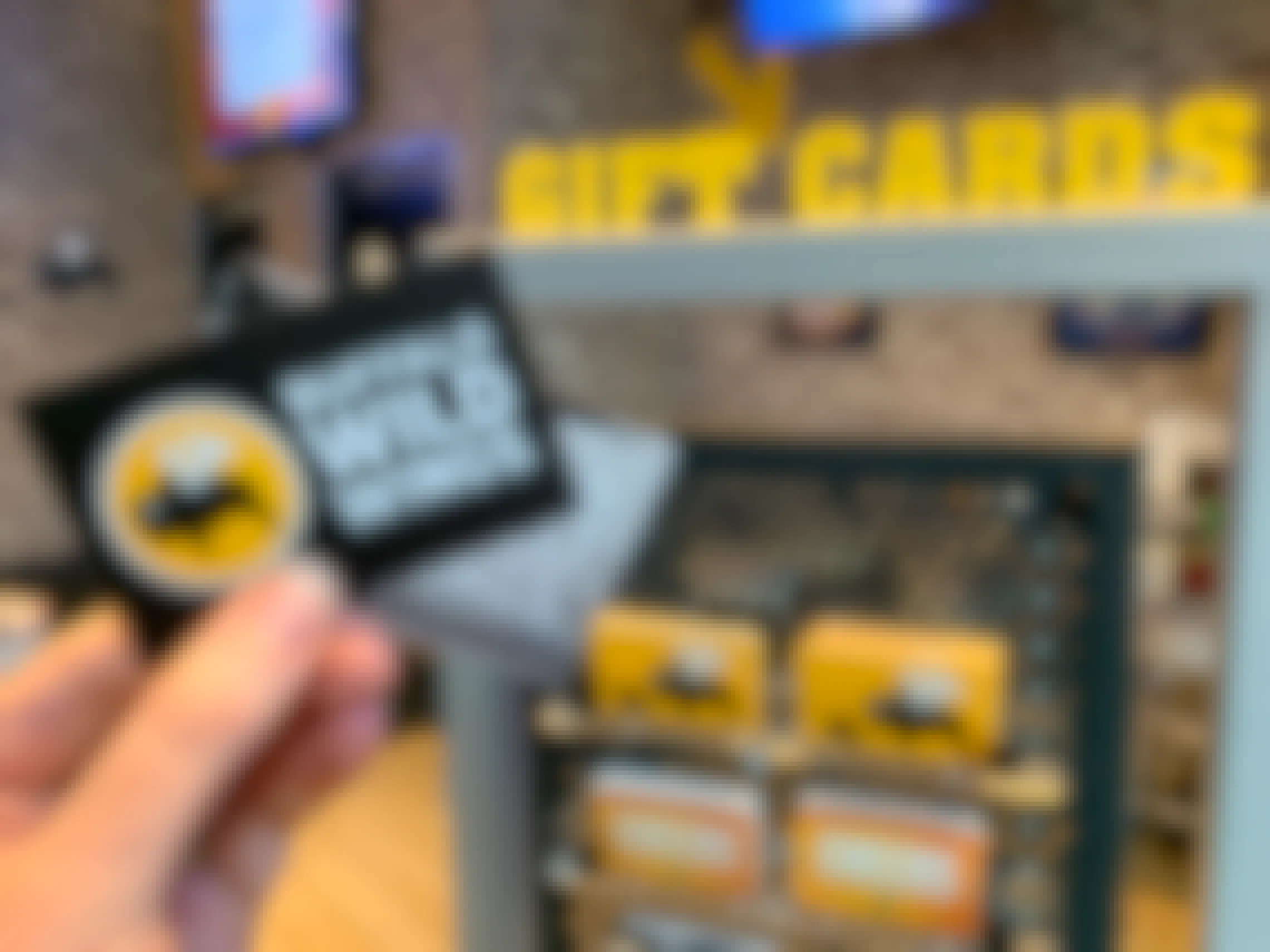 Someone holding two Buffalo Wild Wings gift cards in front of the Gift Card stand inside Buffalo Wild Wings.
