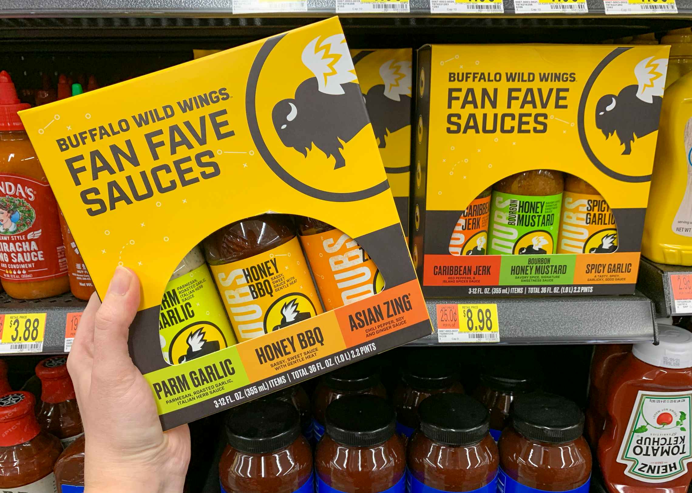 A person holding Buffalo Wild Wings Fan Fave Sauces - a three pack of sauces including Parm Garlic, Honey BBQ, and Asian Zing - in front of a shelf with more of the same product at Walmart.