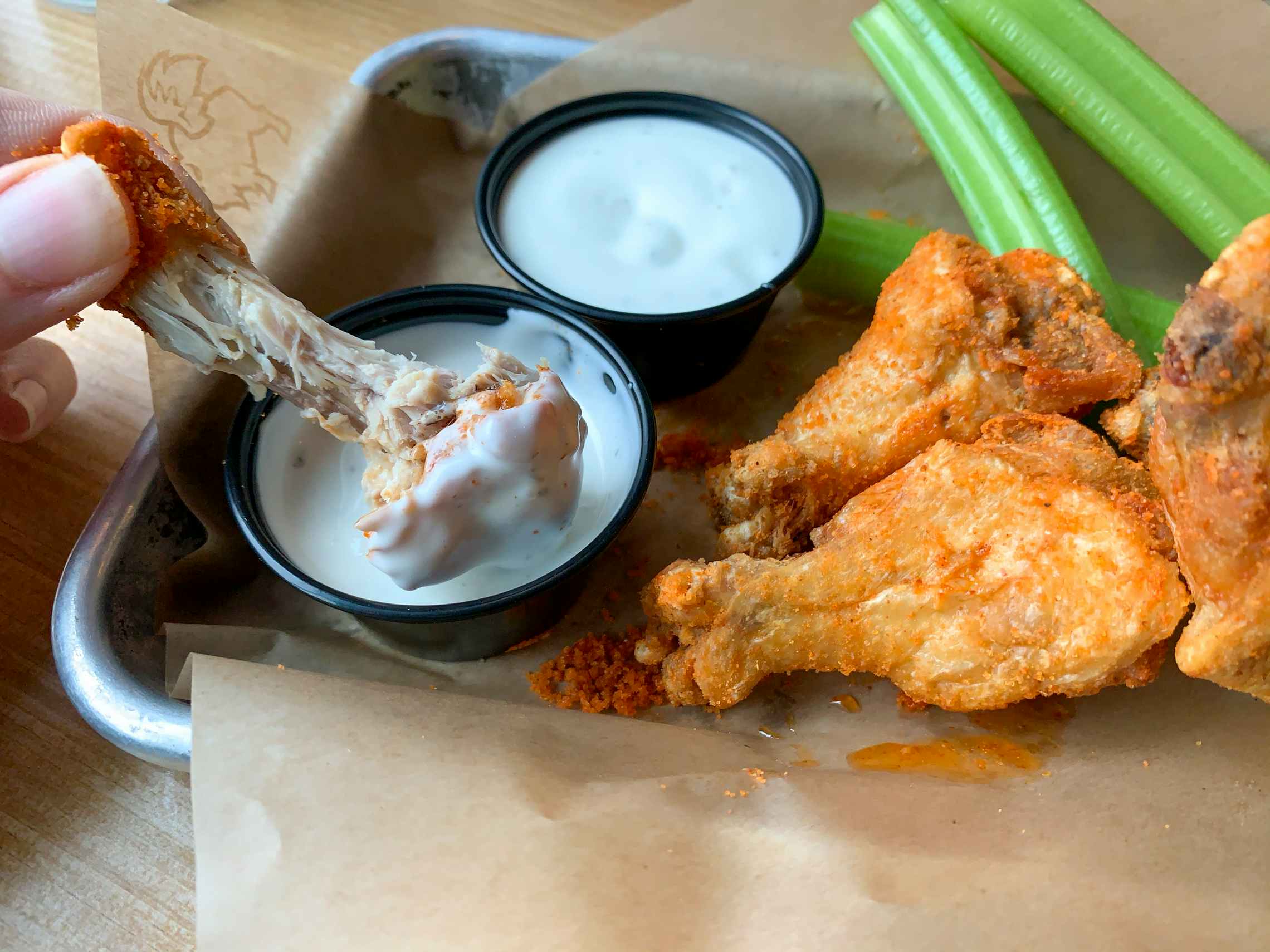 A partially eaten dry rub seasoned traditional buffalo wings being dipped into a ranch dip cup. To the right is a second dip cup, three more wings, and some celery.