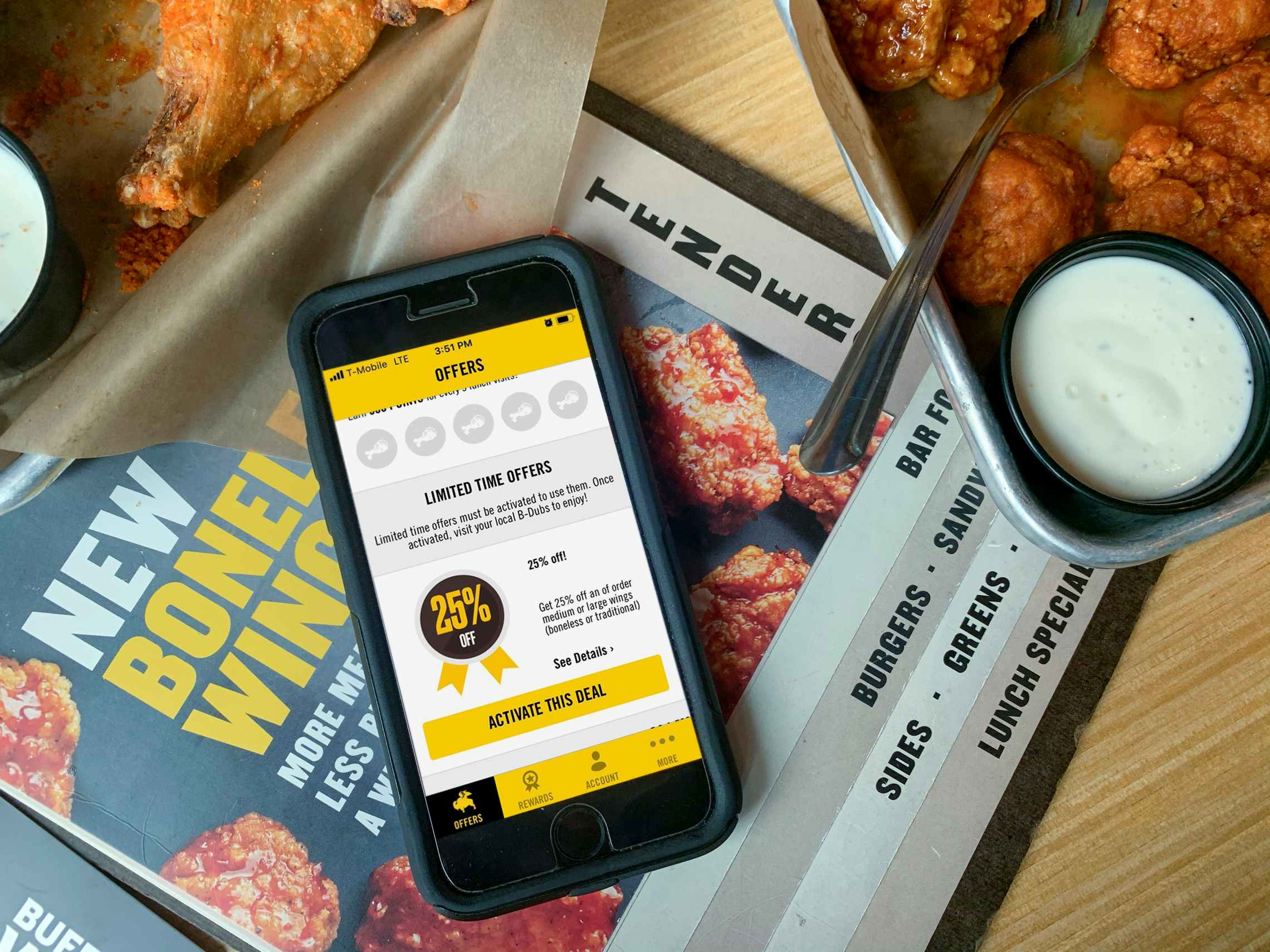 An iPhone sitting on top of a menu on a table, open to the Buffalo Wild Wings app's Offer page. The page shows a 25% off deal that can be activated using the app. To the right of the phone, there is a platter of boneless wings with a fork, and some ranch in a dipping cup.