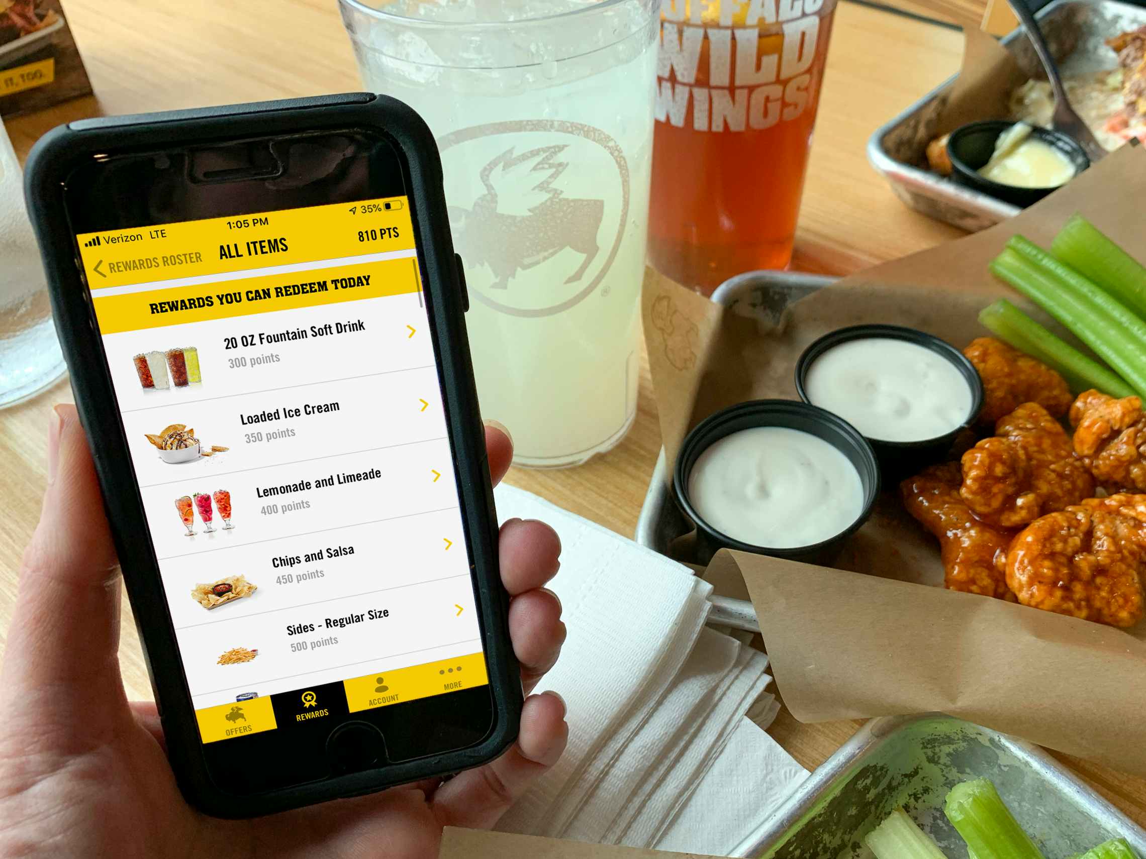 A person holding their iPhone with the Buffalo Wild Wings mobile app open to the Rewards Roster page; the list shows the user's redeemable rewards. On the table in the background, there are two drinks and a platter of breaded boneless wings, two dip cups, and some celery.