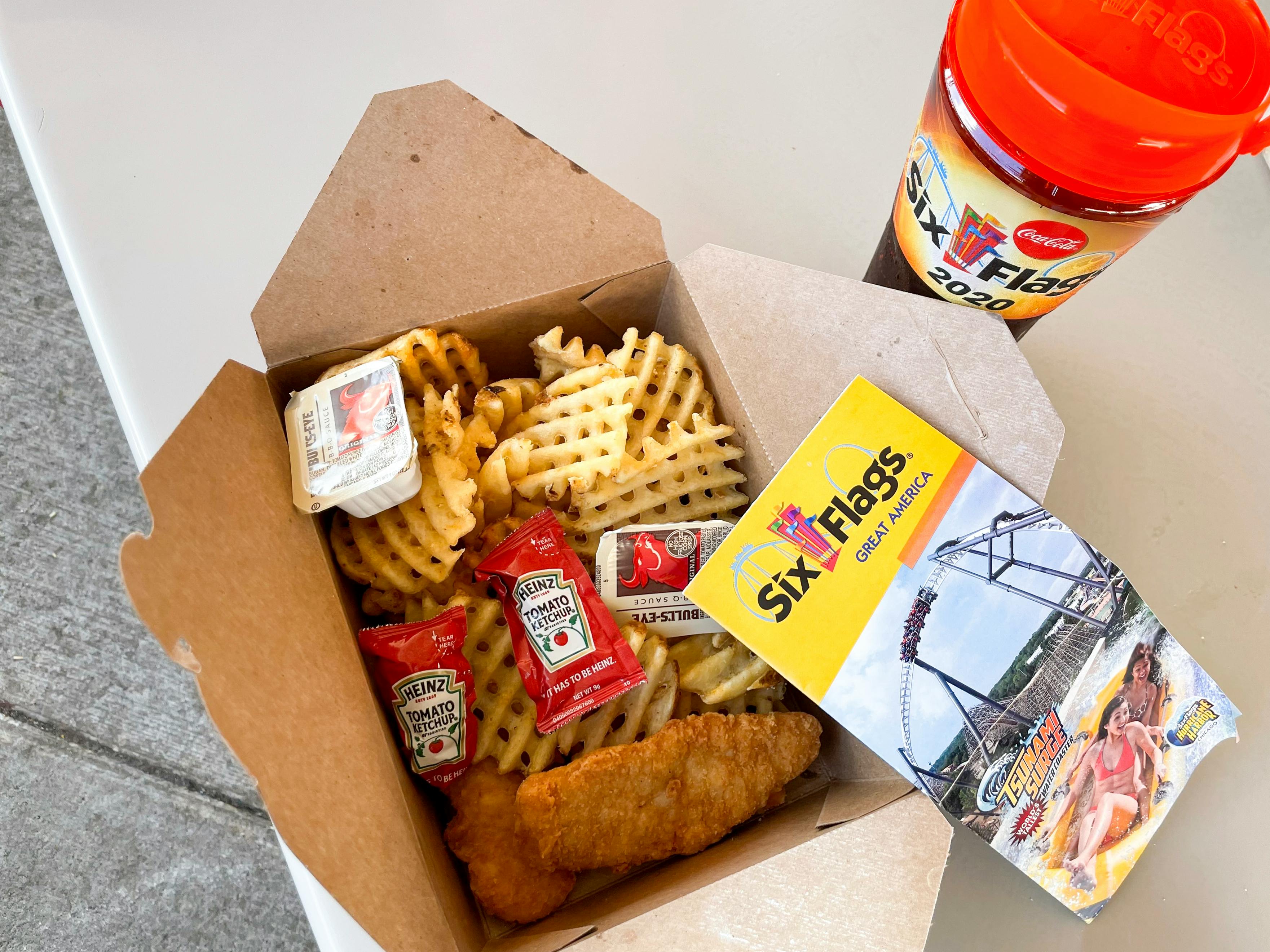 Waffle fries and chicken fingers in a cardboard box next to a refillable soft drink cup and Six Flags theme park map