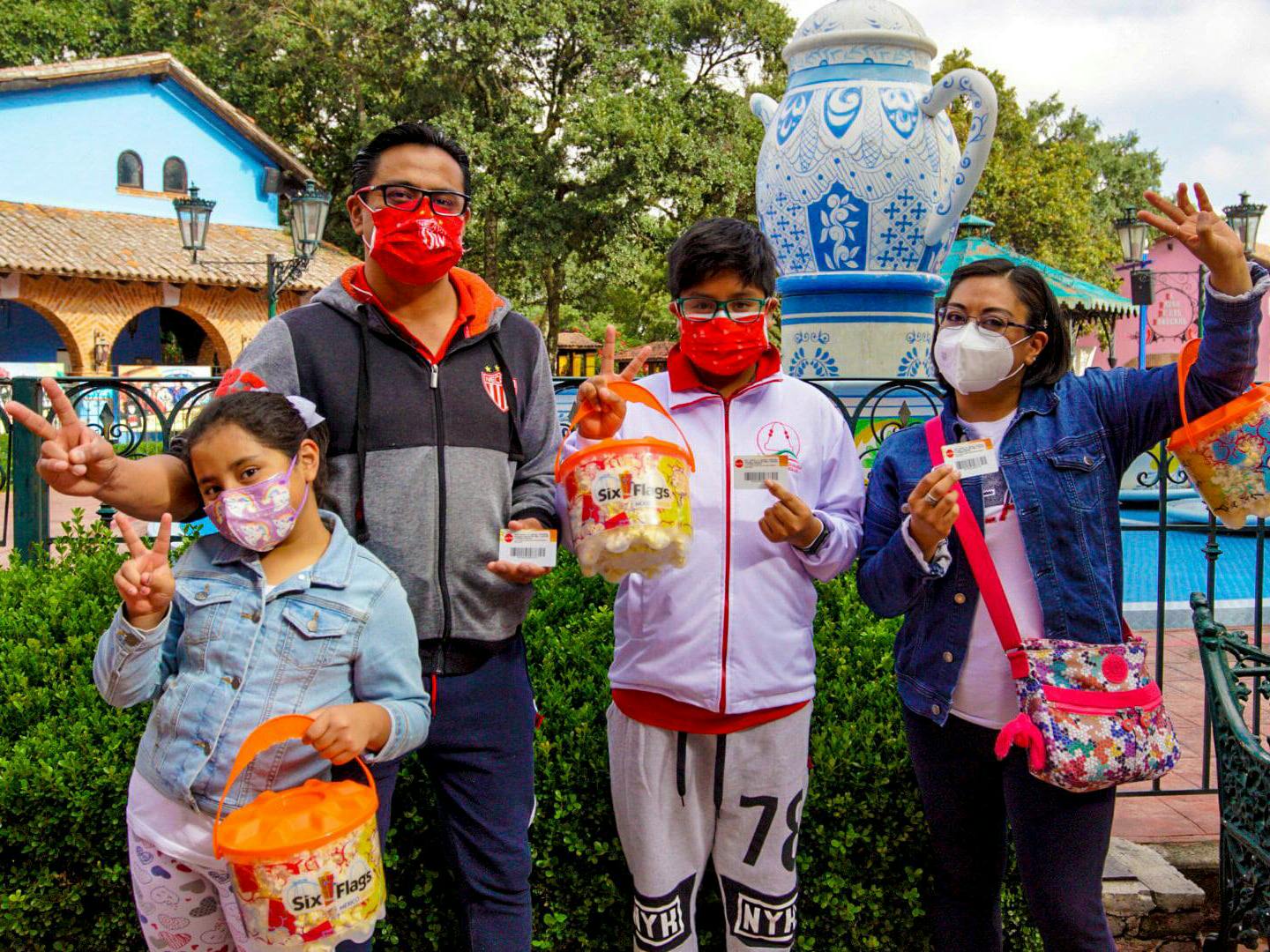 A family of 4 holding up season passes, peace signs, and popcorn, inside a Six Flags amusement park