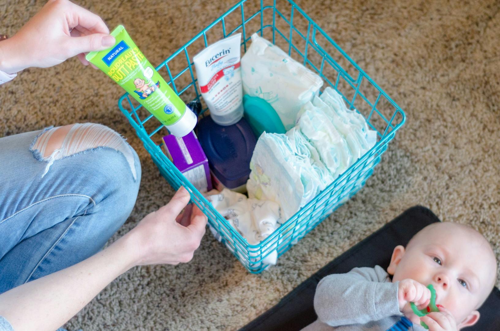 someone putting diaper cream in a basket with diaper supplies in it next to a baby laying on the floor