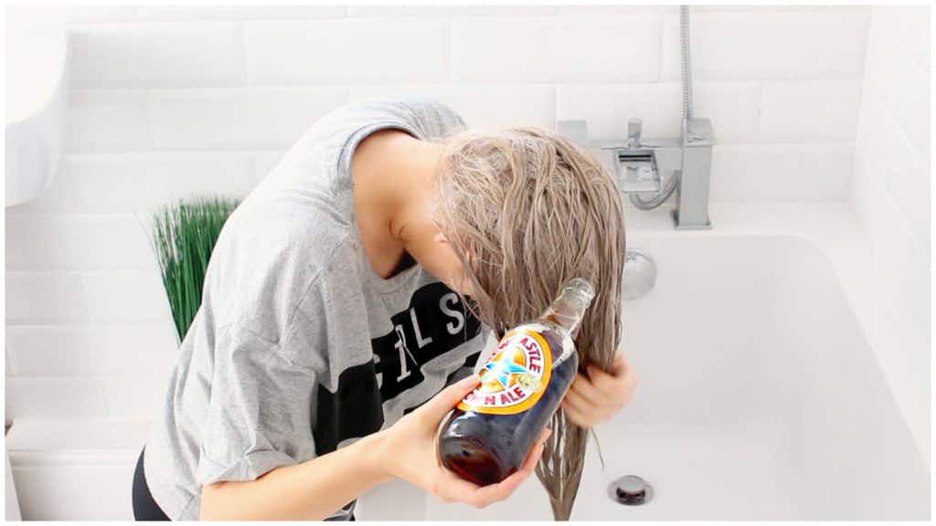 A woman pouring a bottle of beer on her hair as she leans over the tub in a bathroom.