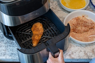 A piece of breaded chicken in an air fryer sitting on a counter next to a bowl of eggs and a bowl of bread crumbs.