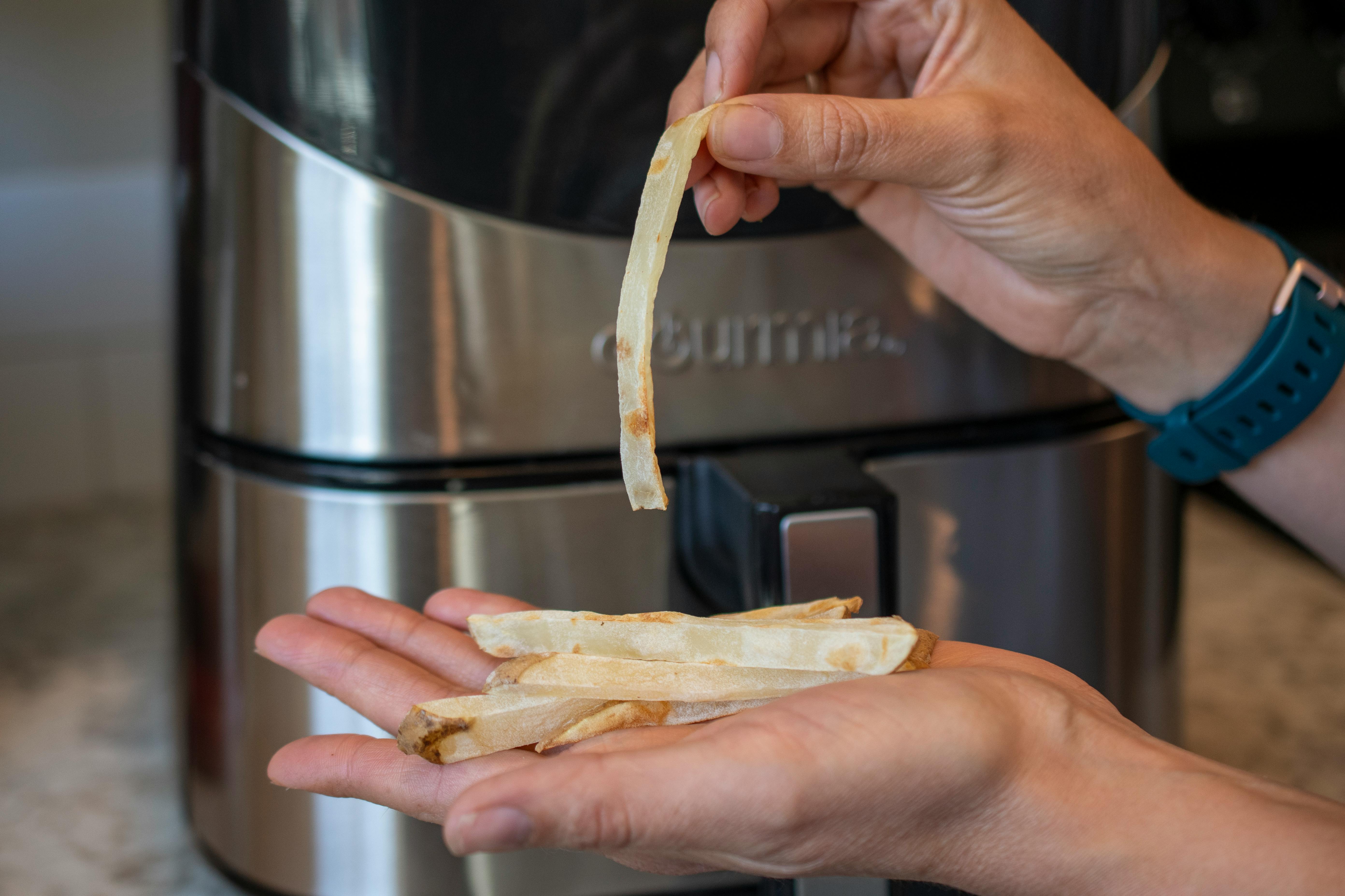 A person's hands holding some limp, dry-looking french fries in front of an air fryer.