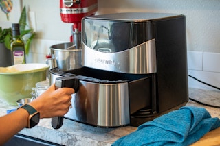 A person's hand pulling the drawer out of air fryer that is sitting on a counter with stuff surrounding it on all sides.