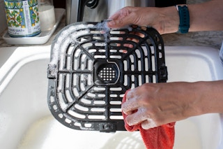 A person hand-washing an air fryer tray in a kitchen sink.