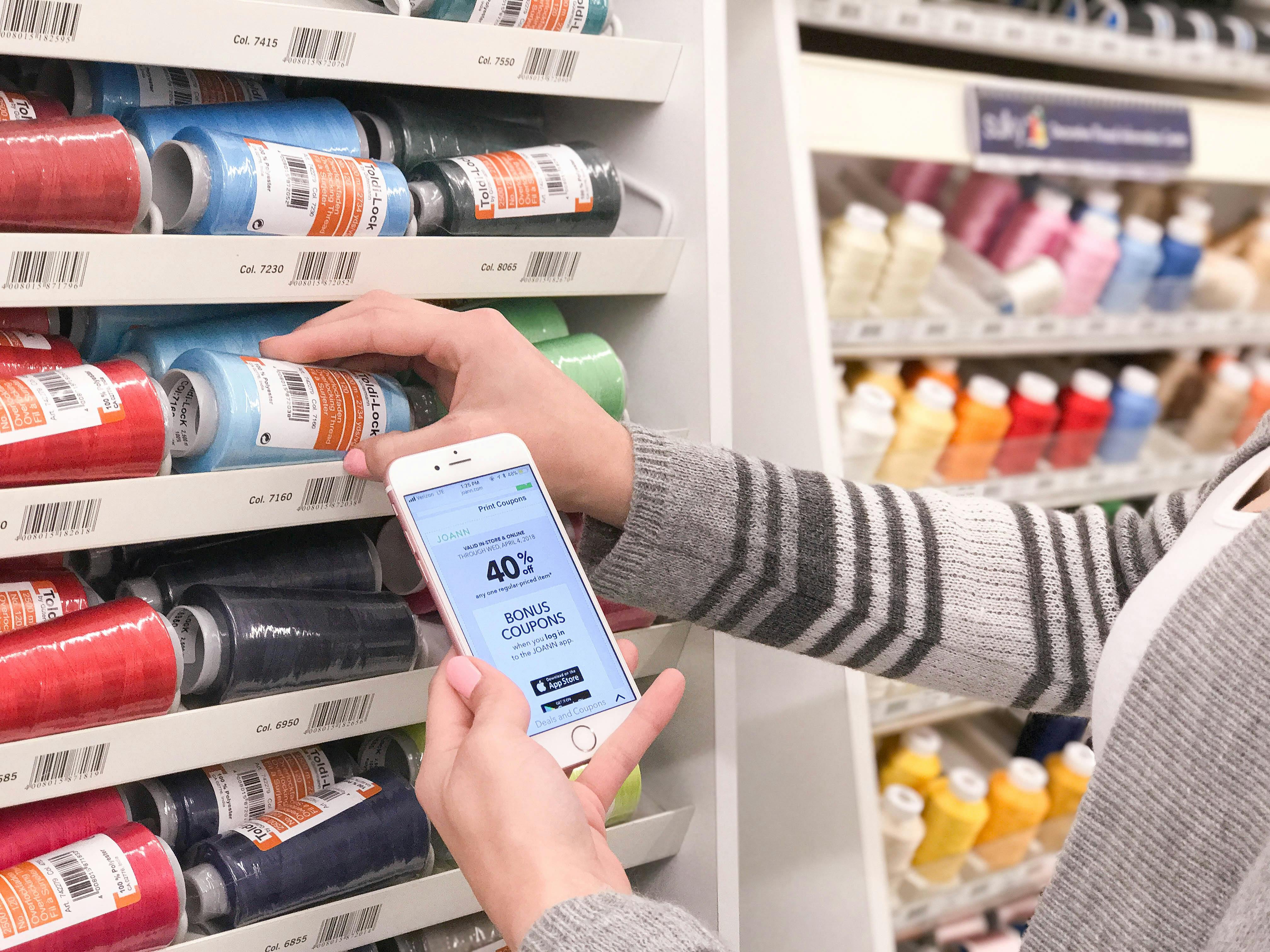 person picking up thread and holding a phone with a 40% coupon on it in front of an aisle of thread at Joann