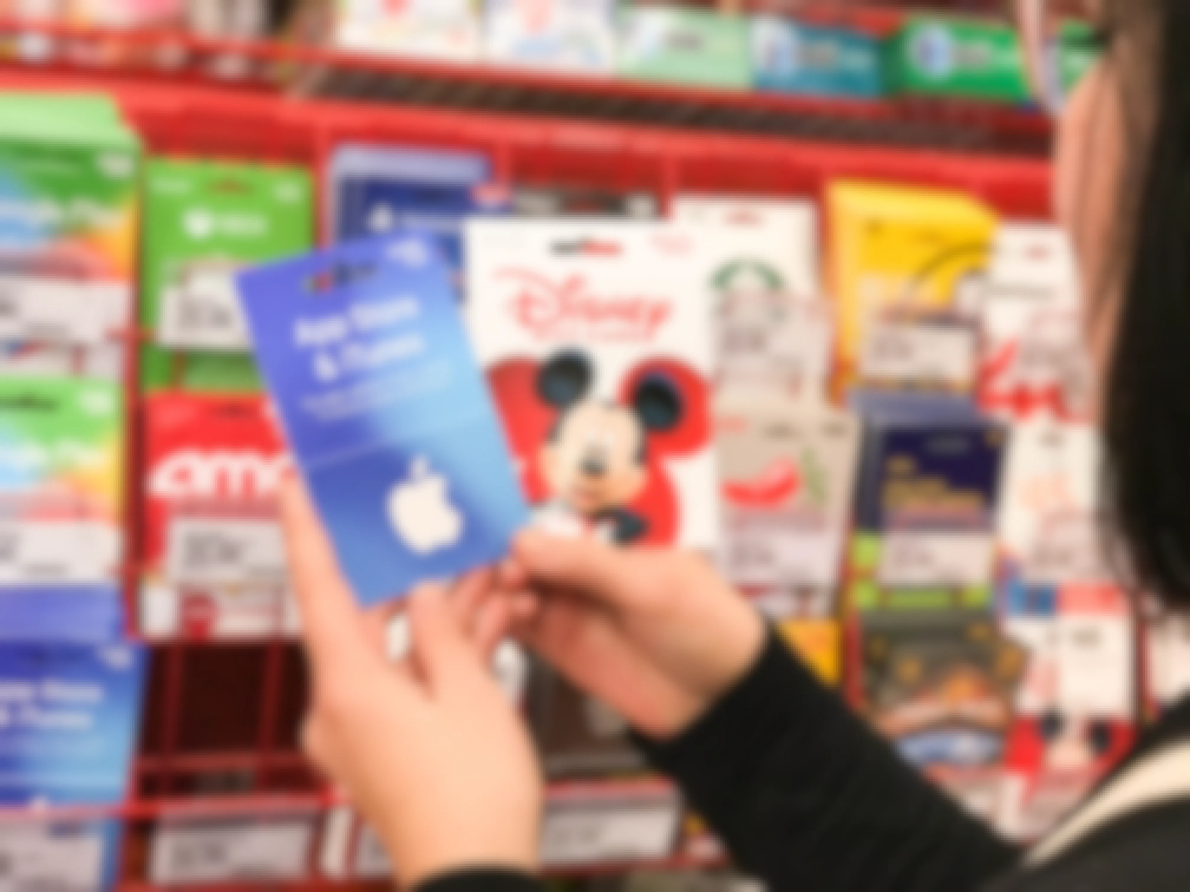 A person holding up two gift cards, one for Disney and one for the Apple App Store & iTunes, in front of the gift card display at Target.