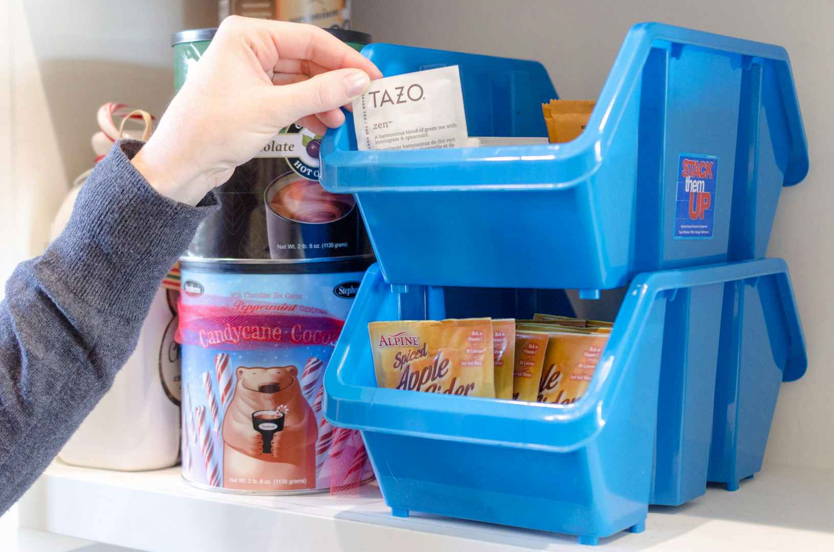 a person taking out a tea packet from stacked up plastic organizing baskets in a pantry 