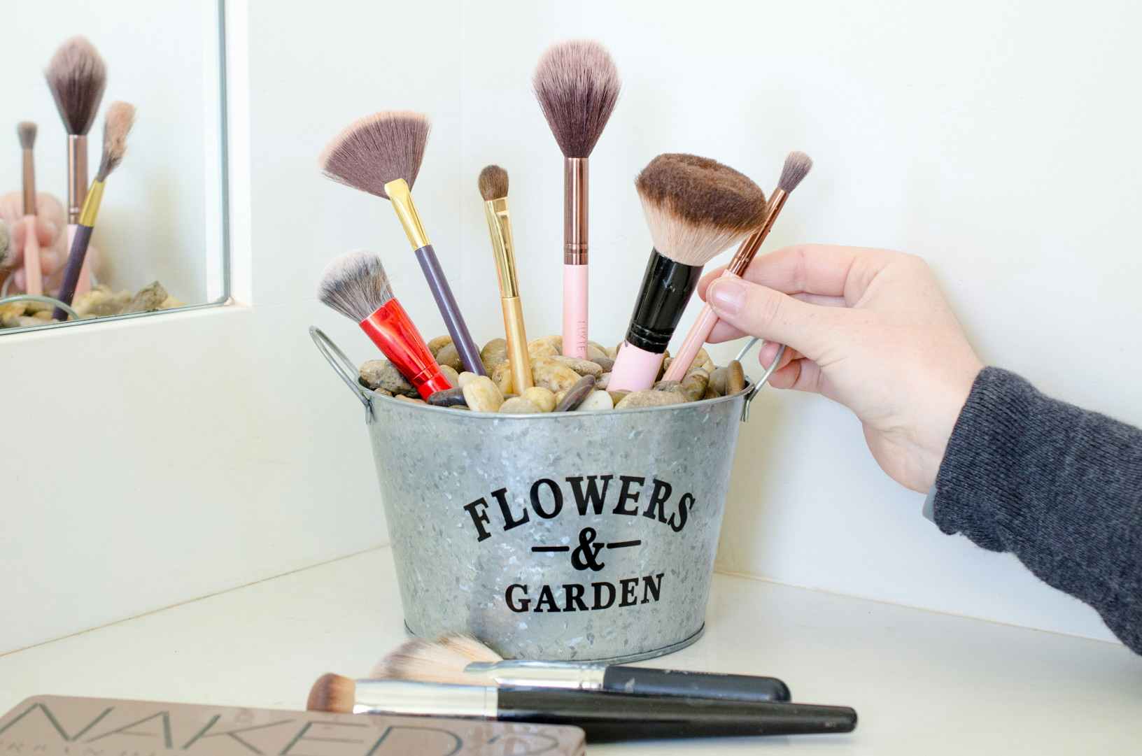 a person placing makeup brushes into a flower tin bucket filled with rocks 