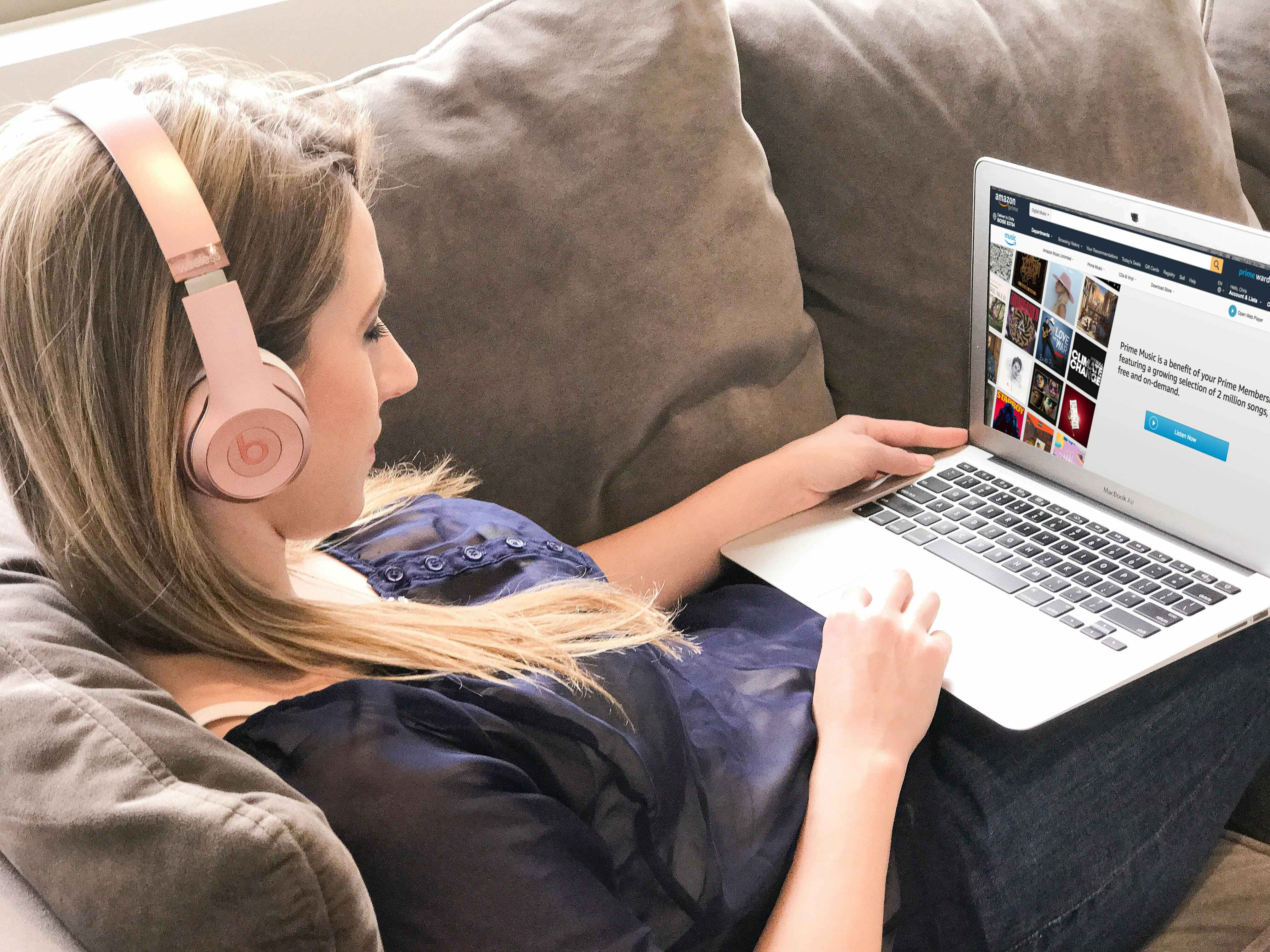 A woman, wearing headphones, sitting on a couch and using her laptop to look at Amazon's Prime Music page on their website.