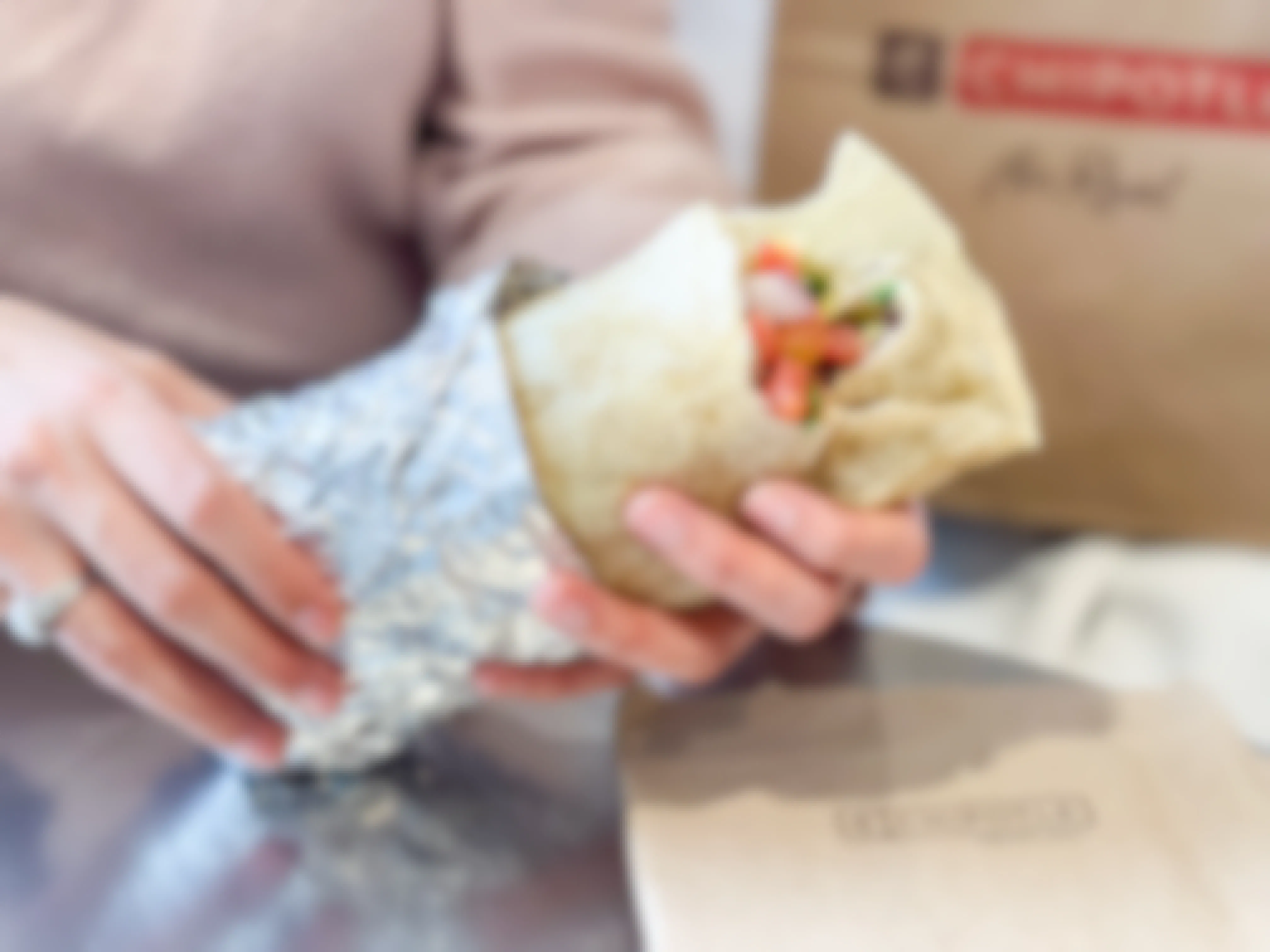 A person's hands holding a burrito that is half wrapped in foil in front of a Chipotle takeout bag.