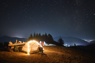 A person sitting outside their tent decorated in lights, looking up at the sky