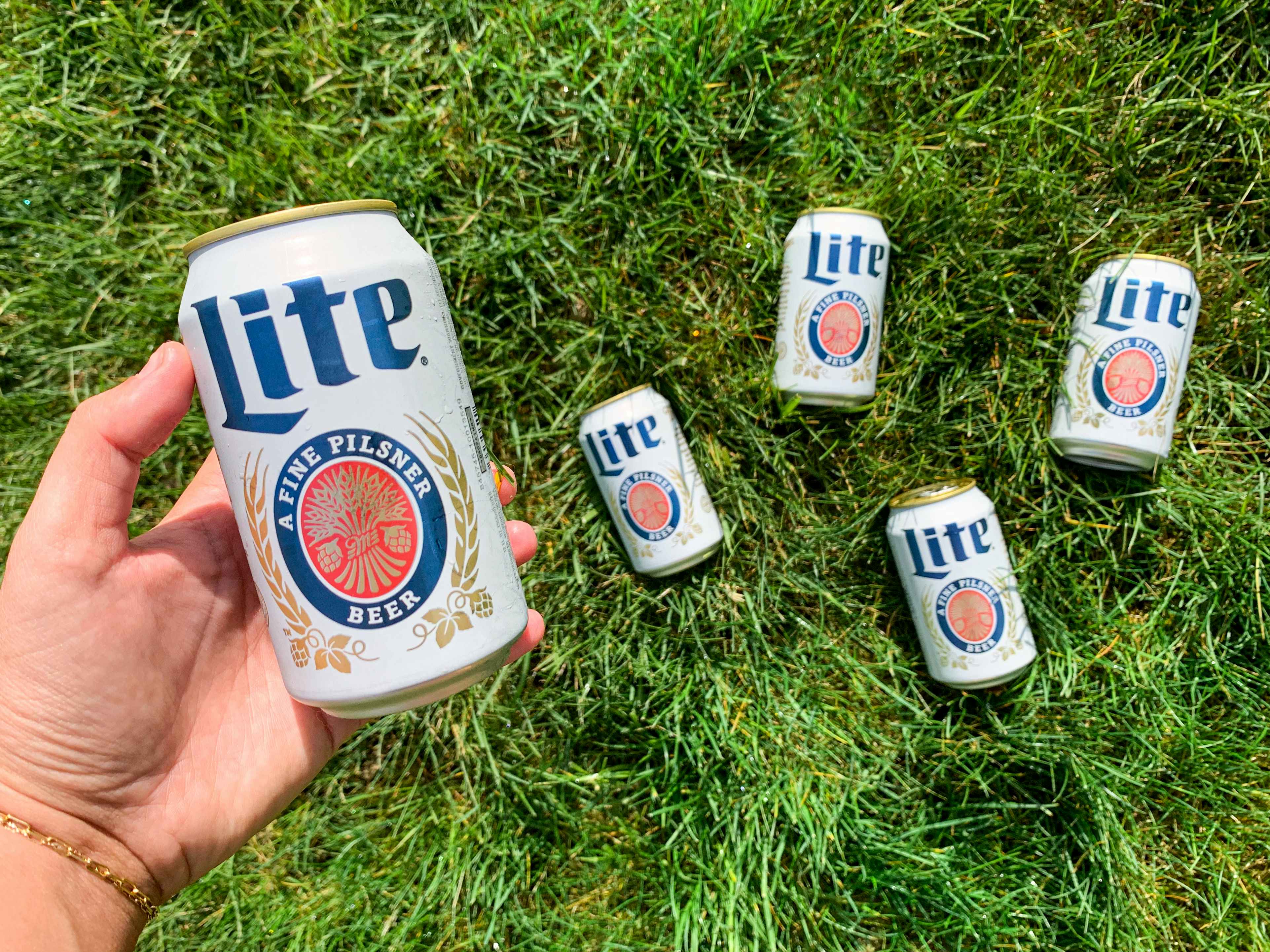 a hand holding miller lite beer with cans in the background laying in the grass