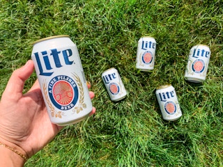 a hand holding miller lite beer with cans in the background laying in the grass