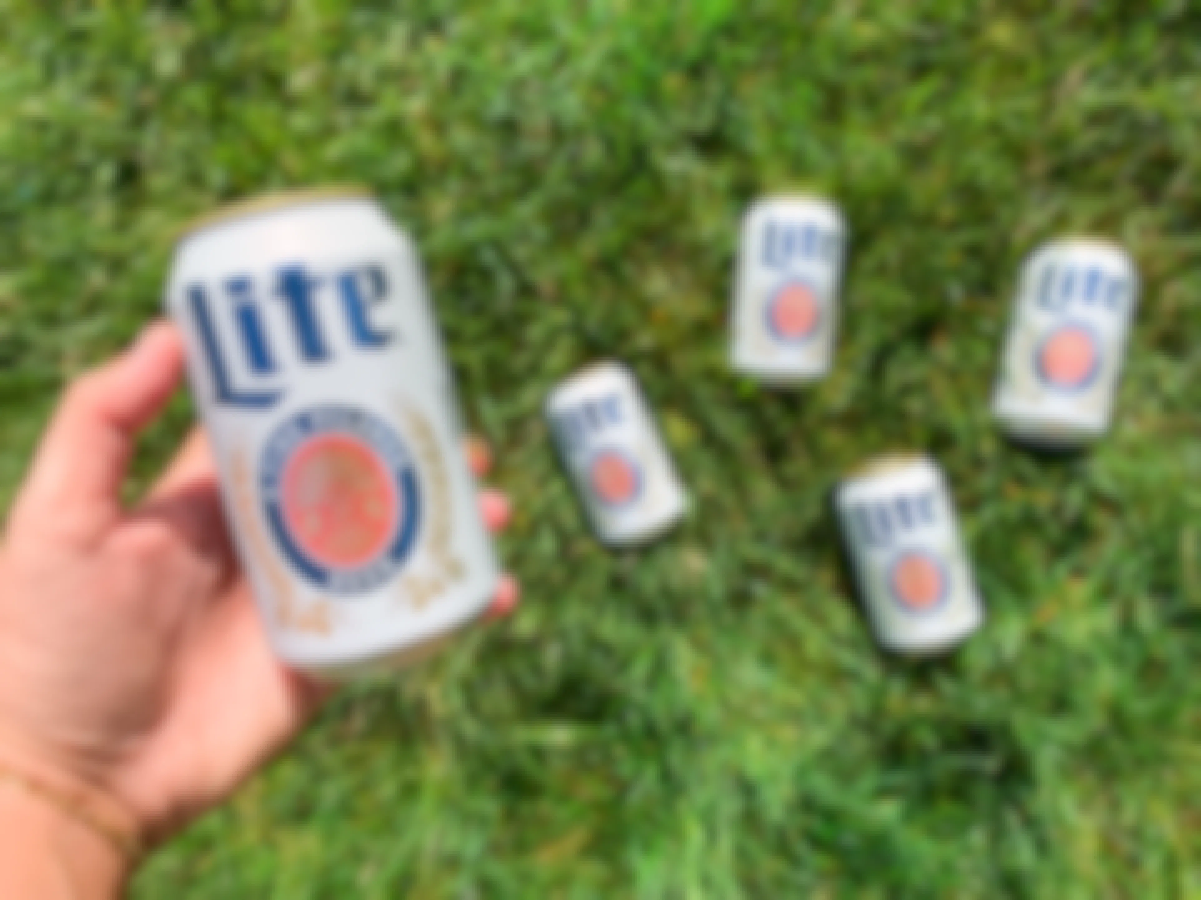 a hand holding miller lite beer with cans in the background laying in the grass