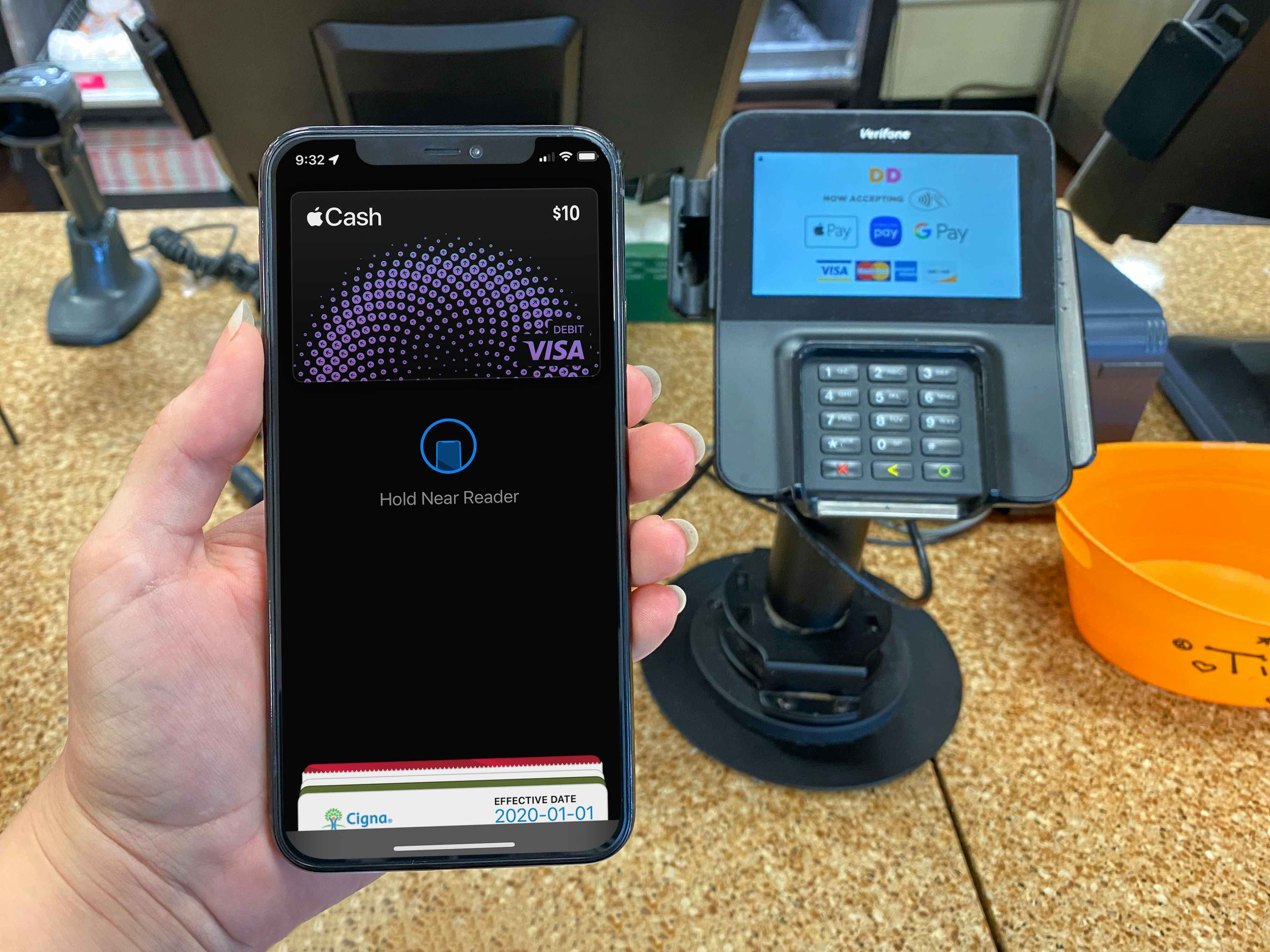 A person's hand holding up an iPhone displaying their Apple Pay page in front of the point-of-sale machine at the counter at Dunkin'.