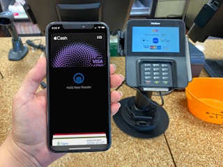 A person's hand holding up an iPhone displaying their Apple Pay page in front of the point-of-sale machine at the counter at Dunkin'.