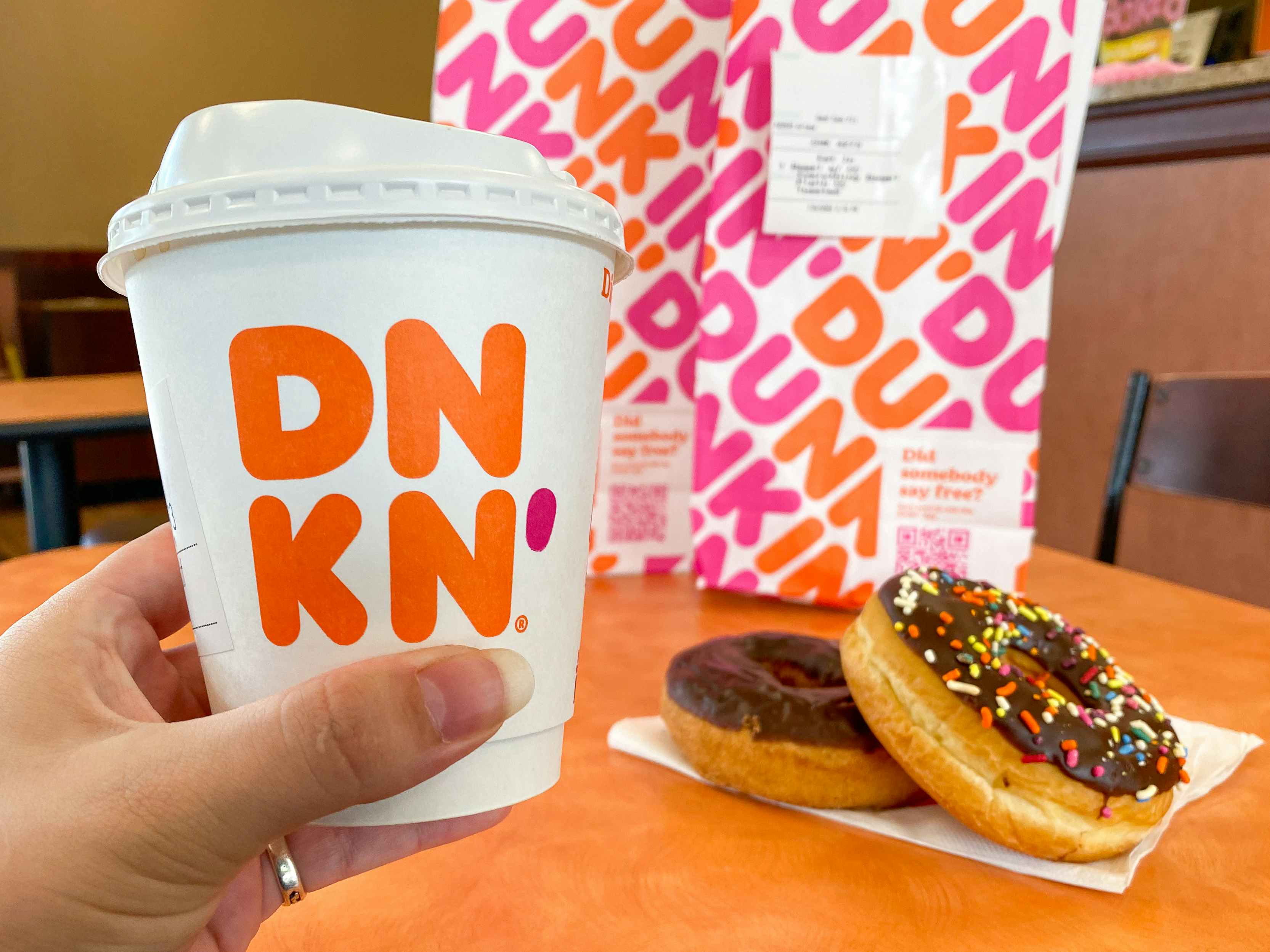 A person's hand holding up a small Dunkin coffee in front of some donuts and a bag sitting on a table inside Dunkin.