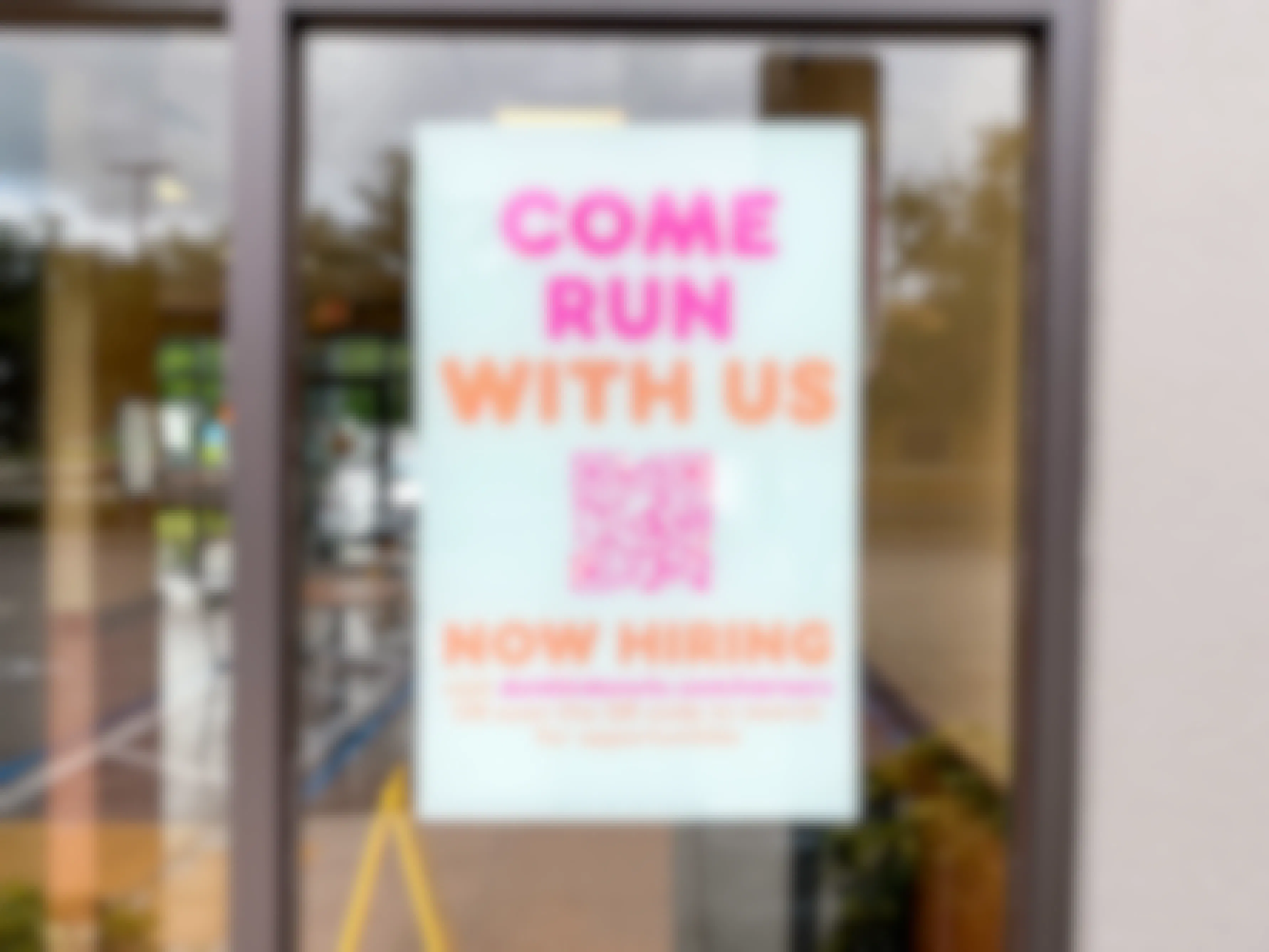 A sign in the window at Dunkin that says "Come run with us" with a QR code to apply for a position at Dunkin.