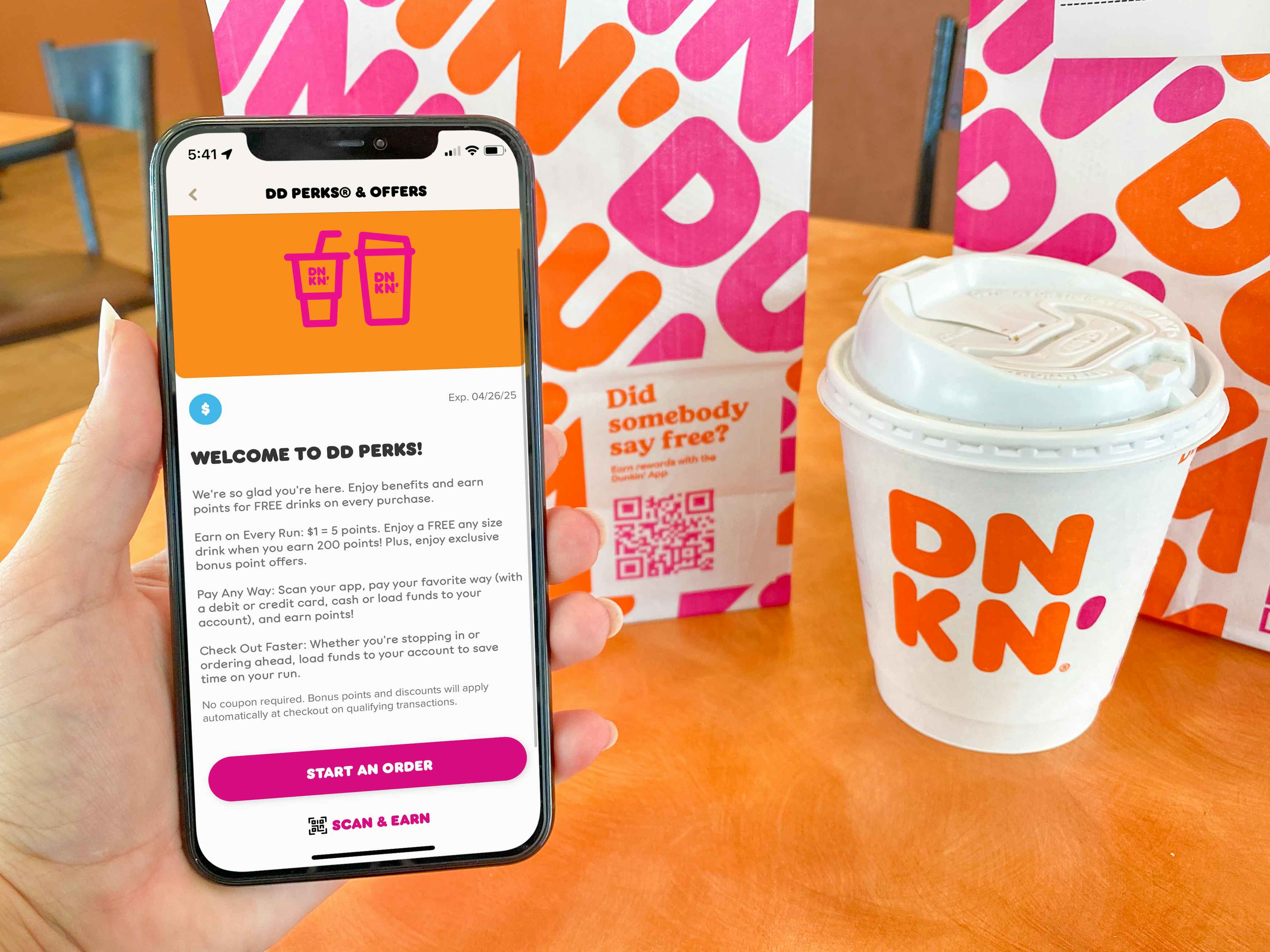 person holding iPhone with dunkin' app DDPerks near bag and coffee