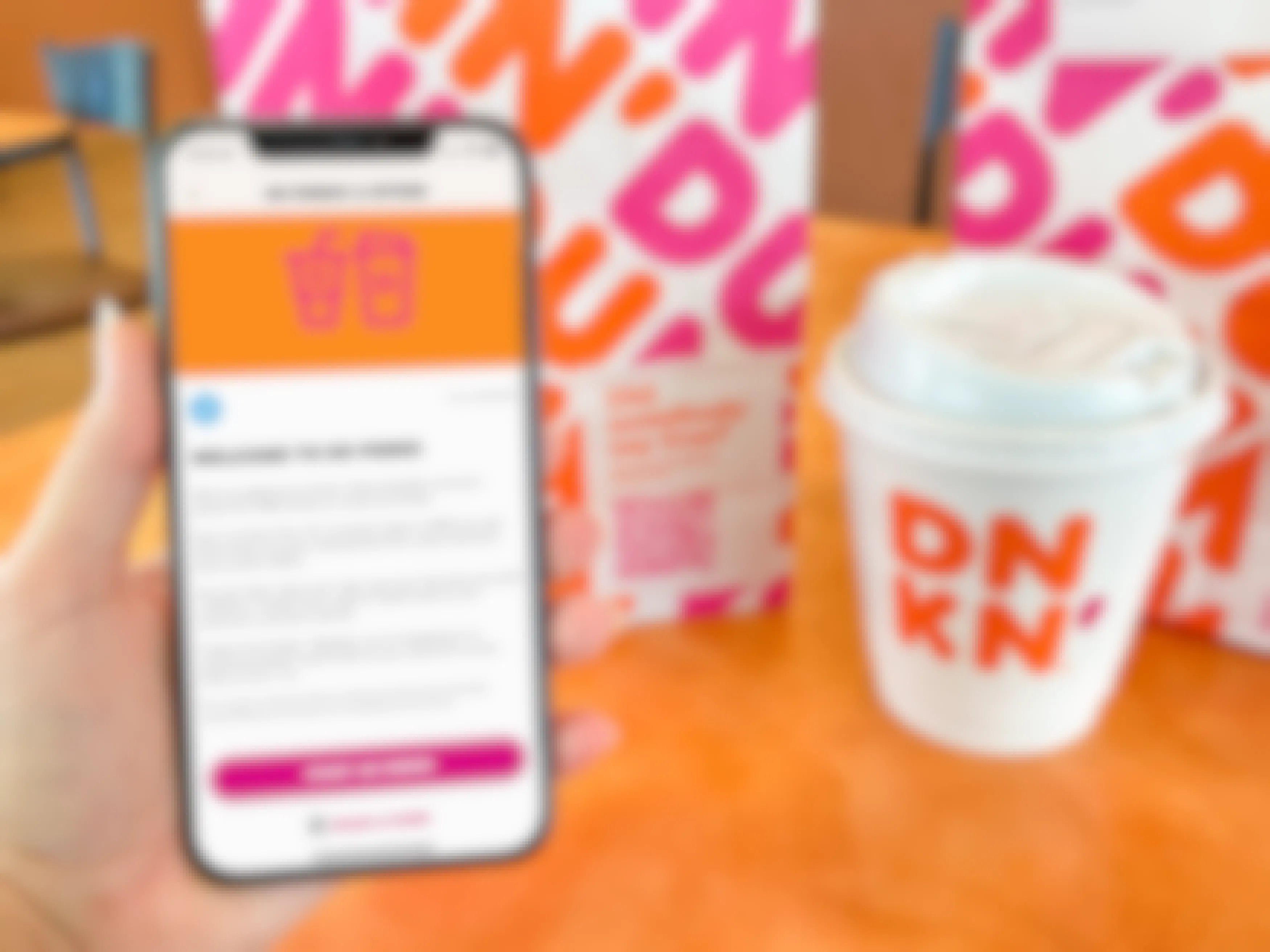 A person's hand holding an iPhone displaying the welcome to DDPerks page on the Dunkin app next to a small coffee and a Dunkin bag inside Dunkin.