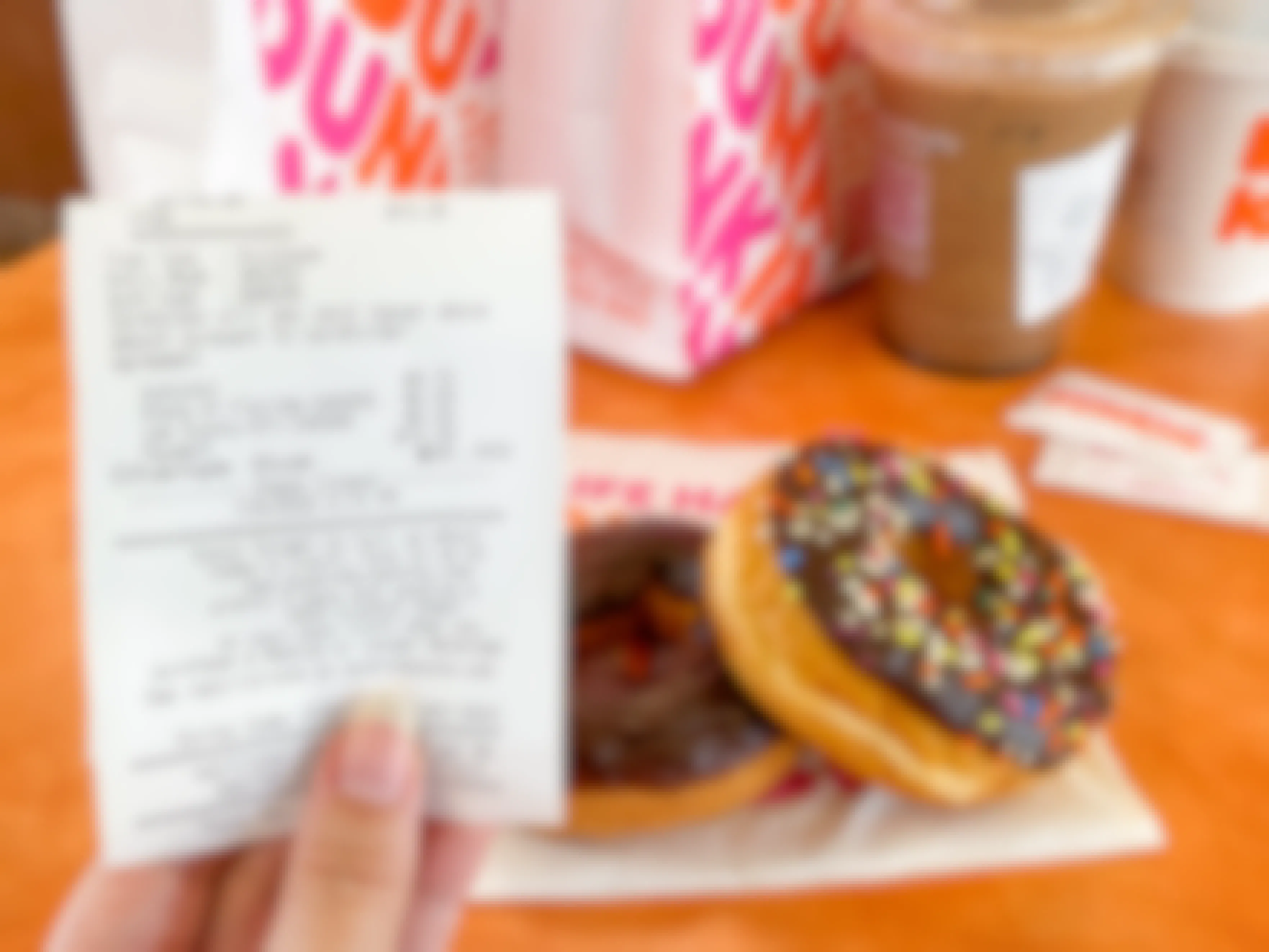 A person's hand holding a receipt offering a free donut for taking the survey printed on the bottom with some donuts, two coffees, some sugar packets, and Dunkin bags on the table in the background.