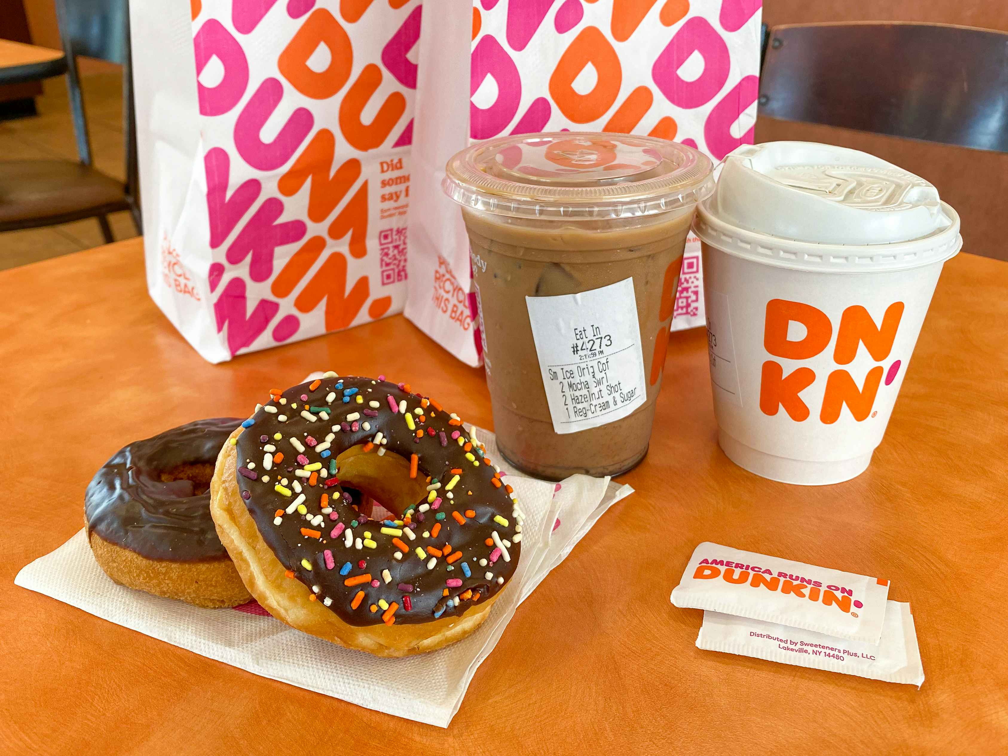 Two donuts on a napkin, some sugar packets, two coffees, and two Dunkin bags sitting on a table inside Dunkin.