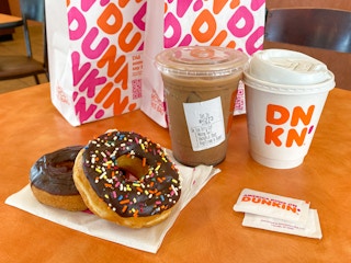 Two donuts on a napkin, some sugar packets, two coffees, and two Dunkin bags sitting on a table inside Dunkin.
