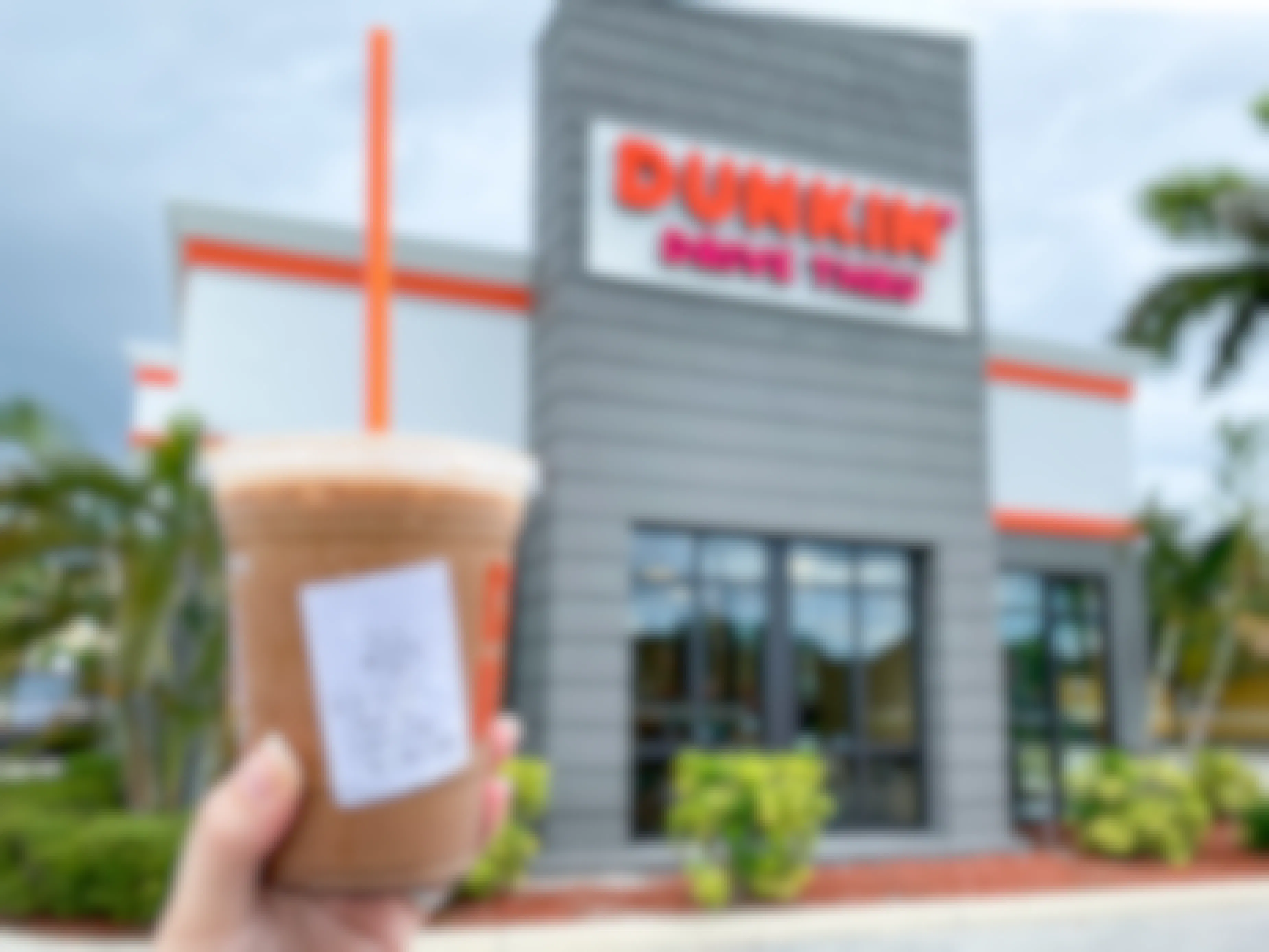 A person's hand holding up a specialty iced Dunkin Donuts coffee outside of a Dunkin restaurant.