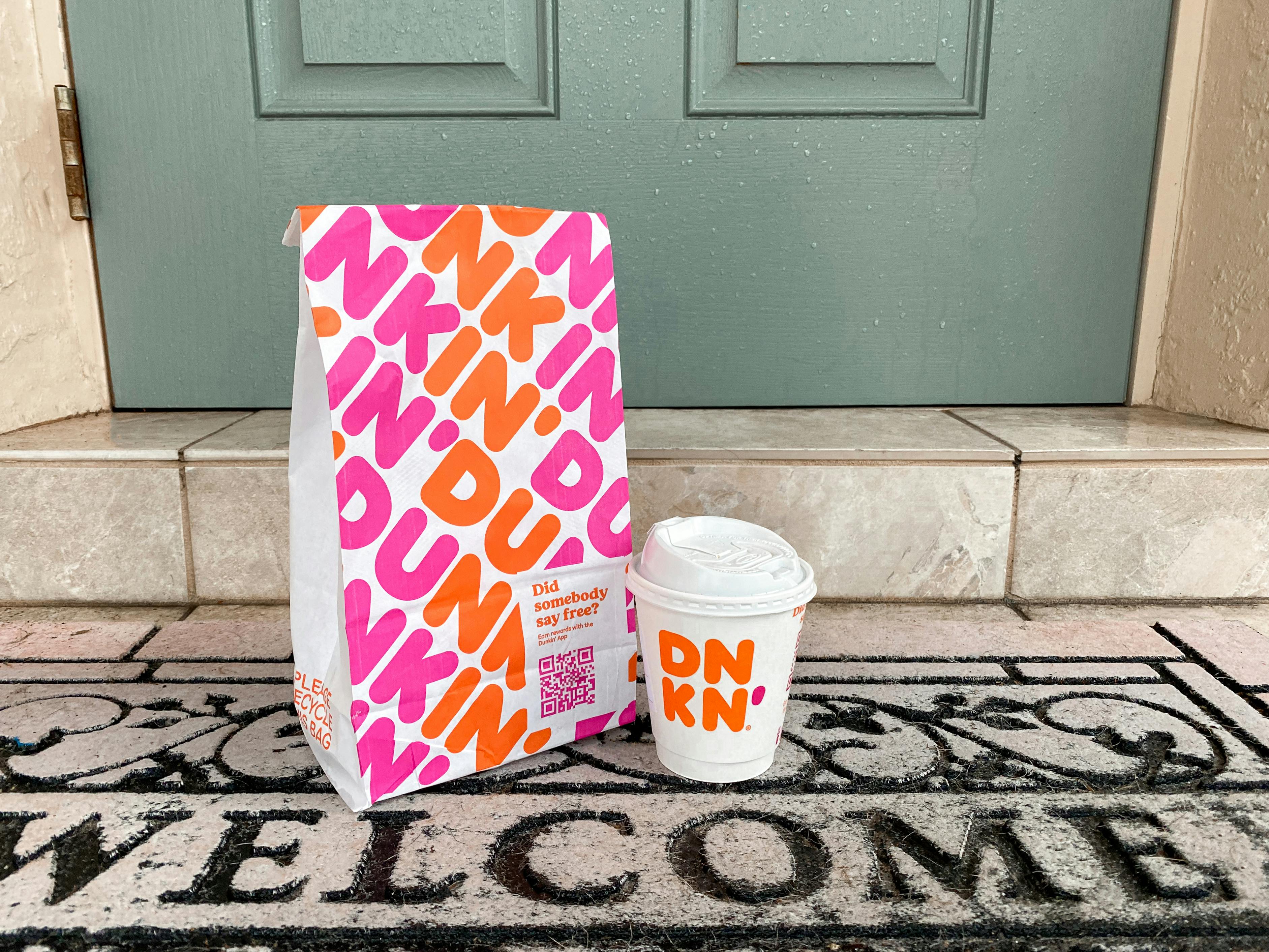 A Dunkin takeout bag and small coffee sitting on a welcome mat outside of someone's front door.