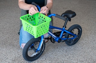 A person tying a green basket to the handlebars of a child's bike