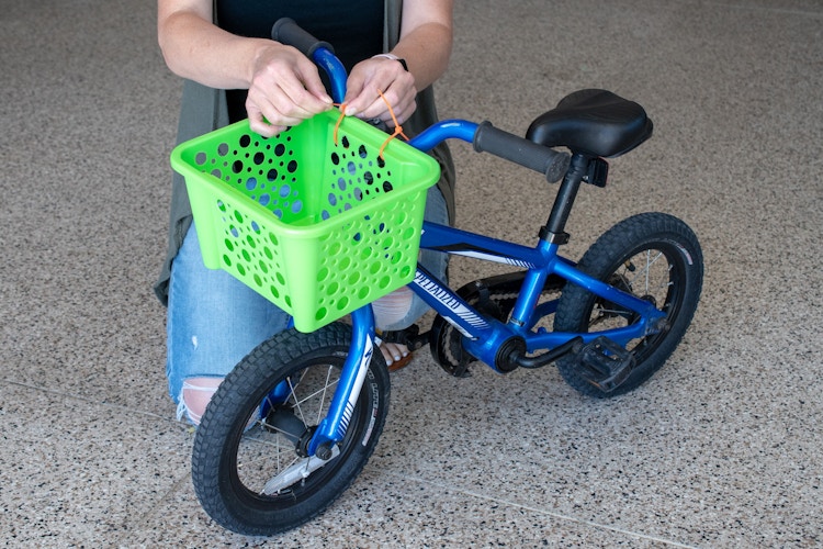 A person tying a green basket to the handlebars of a child's bike