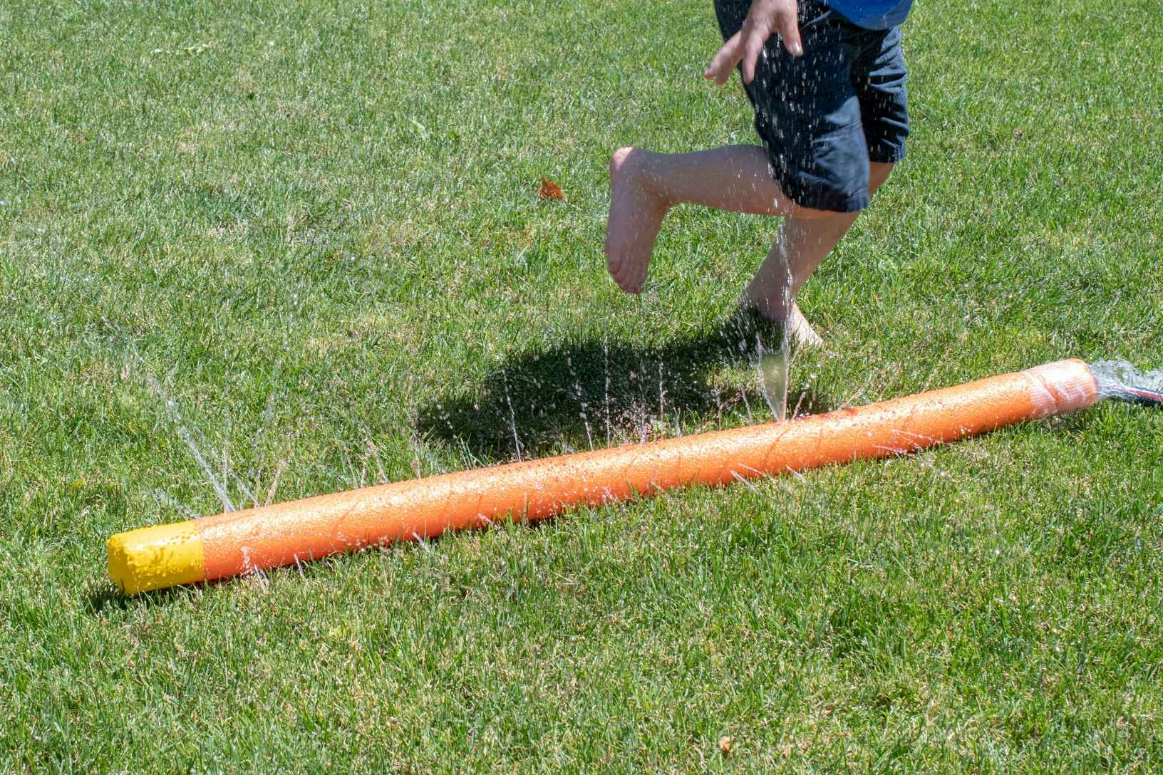 A child playing around a DIY pool noodle sprinkler that is attached to a hose on a lawn.