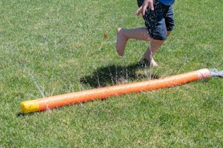 A child playing around a DIY pool noodle sprinkler that is attached to a hose on a lawn.