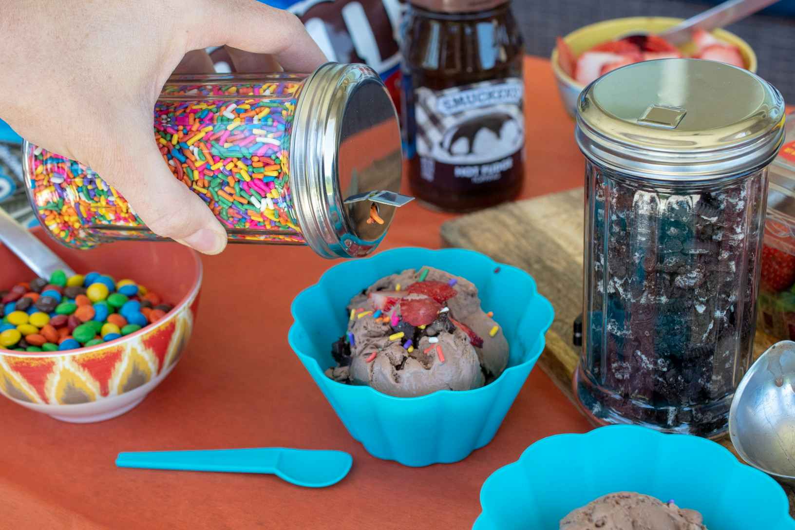 A person's hand pouring a jar of sprinkles onto a small bowl of ice cream on a table next to more ice cream sundae ingredients.