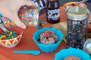 A person's hand pouring a jar of sprinkles onto a small bowl of ice cream on a table next to more ice cream sundae ingredients.